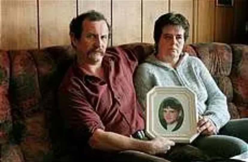 To personer sitter tett sammen i en sofa, og den ene holder en innrammet oval portrett­ramme med et bilde plassert i midten. Bakgrunnen viser trepanelt vegg og mønstret sofatrekk.