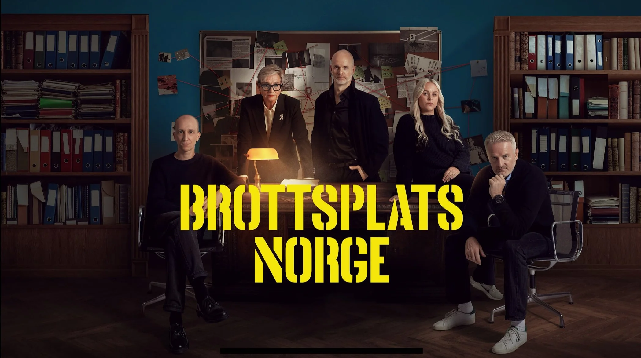 Fem personer sitter og står rundt et skrivebord i et mørkt kontorinteriør. På veggen bak henger et stort brett fylt med notater, papirer og bilder festet med tegnestifter og klyper. Rommet er omgitt av høye bokhyller fulle av permer og mapper.