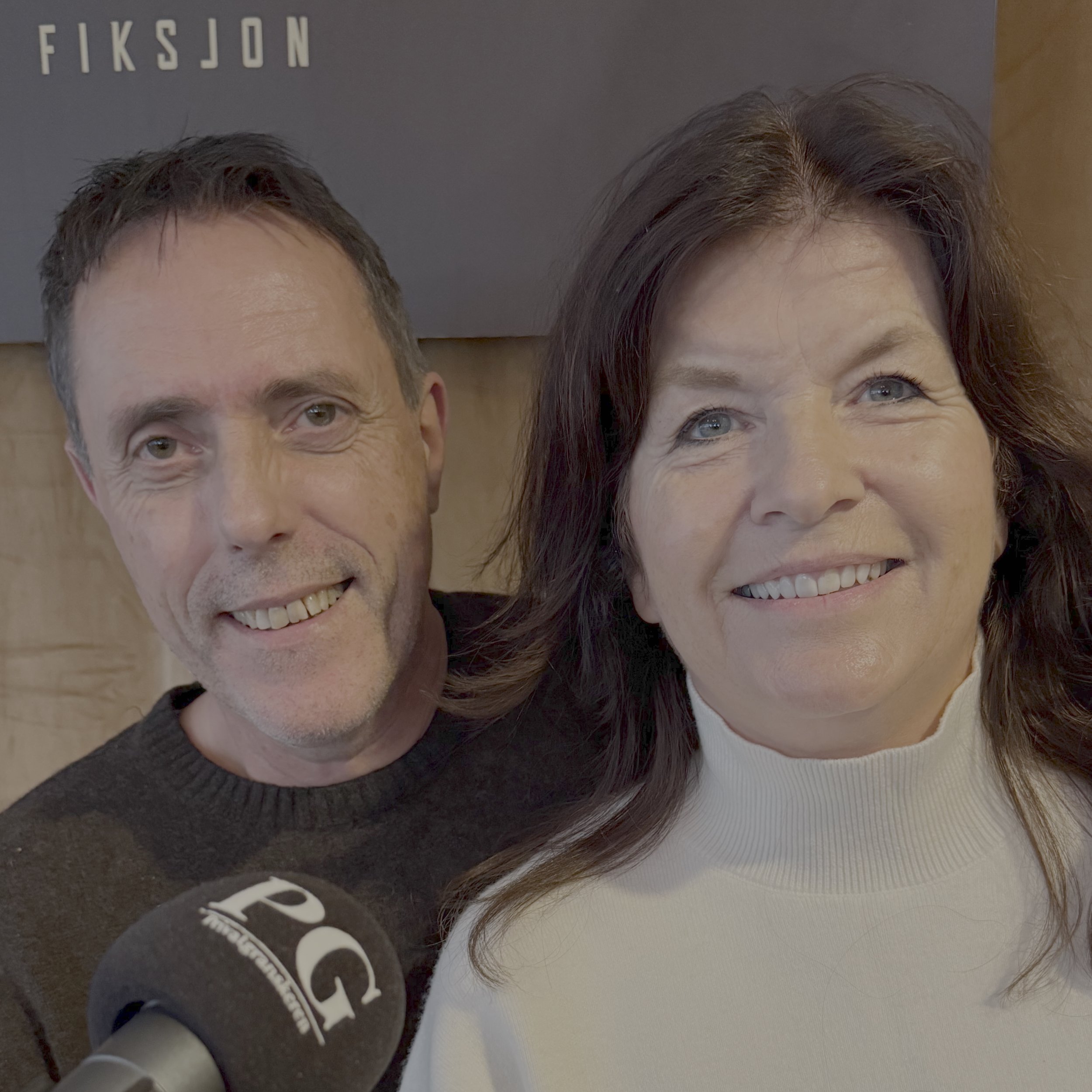 To personer sitter tett foran en PG‑merket podkastmikrofon. Den ene personen har mørkt hår og mørk genser, mens den andre har langt mørkt hår og en lys, høyhalset genser. Bak dem henger et mørkt stoffbanner med delvis synlig tekst.