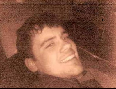 Utsnitt av en person sittende i et mørkt rom, med synlige konturer av hode og overkropp. Bildet har kraftig kornete tekstur og sepia‑aktig fargetone, med uklare detaljer i bakgrunnen.