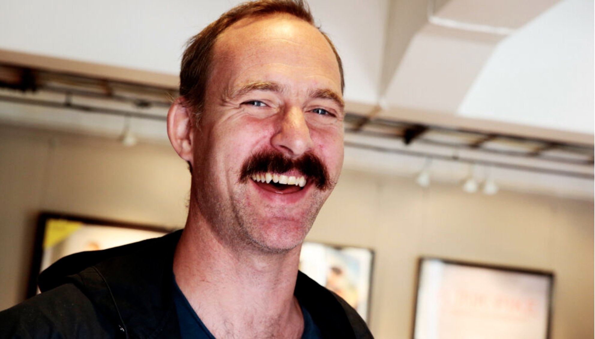 Person med bart står innendørs i et galleri  eller utstillingsmiljø, med plakater og kunstverk synlig i bakgrunnen under takmonterte spotlights.