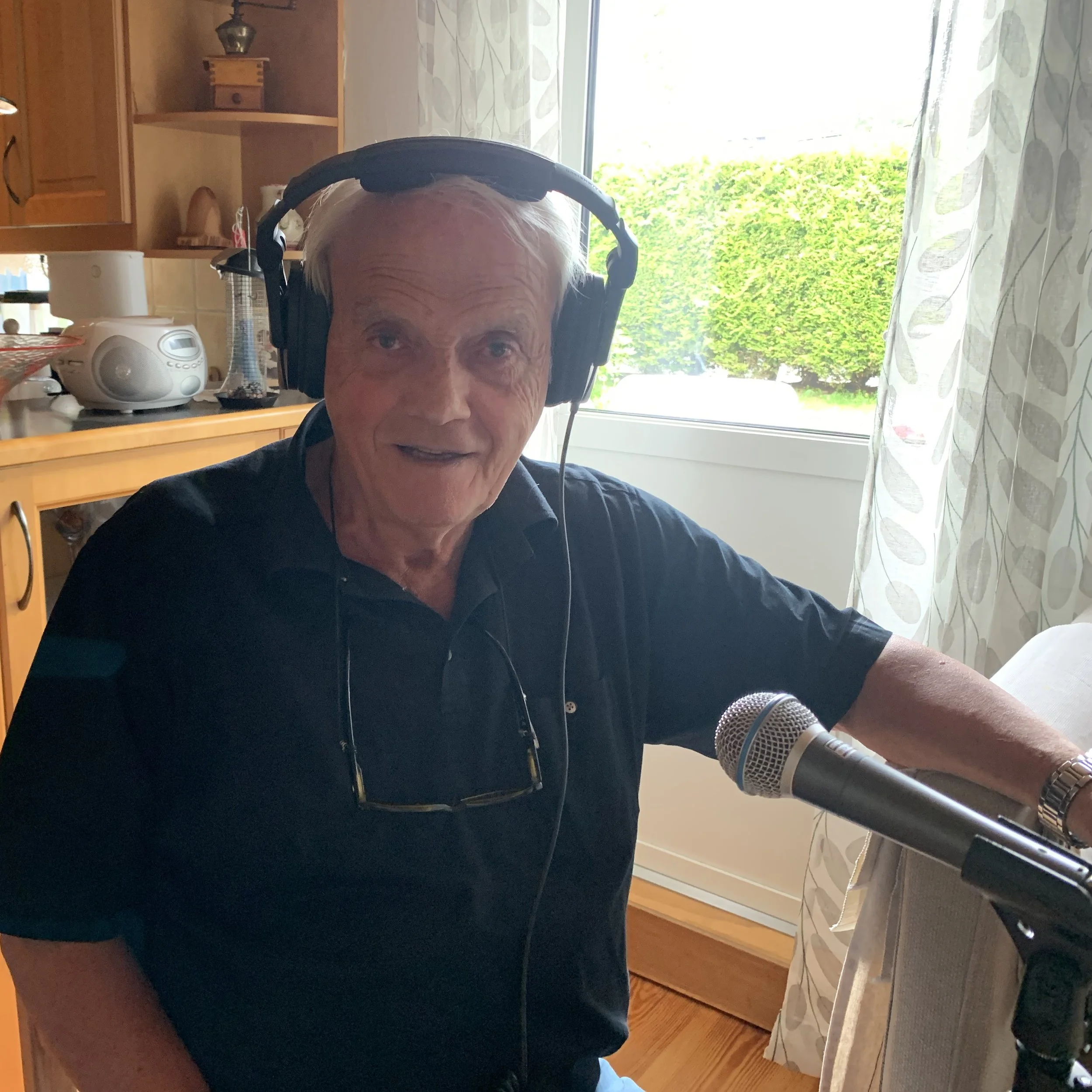 Person i et opptaksoppsett for podcast, sittende ved et bord med hodetelefoner og mikrofon. Bildet er tatt i et kjøkkenmiljø med vindu, gardiner og trehyller i bakgrunnen.