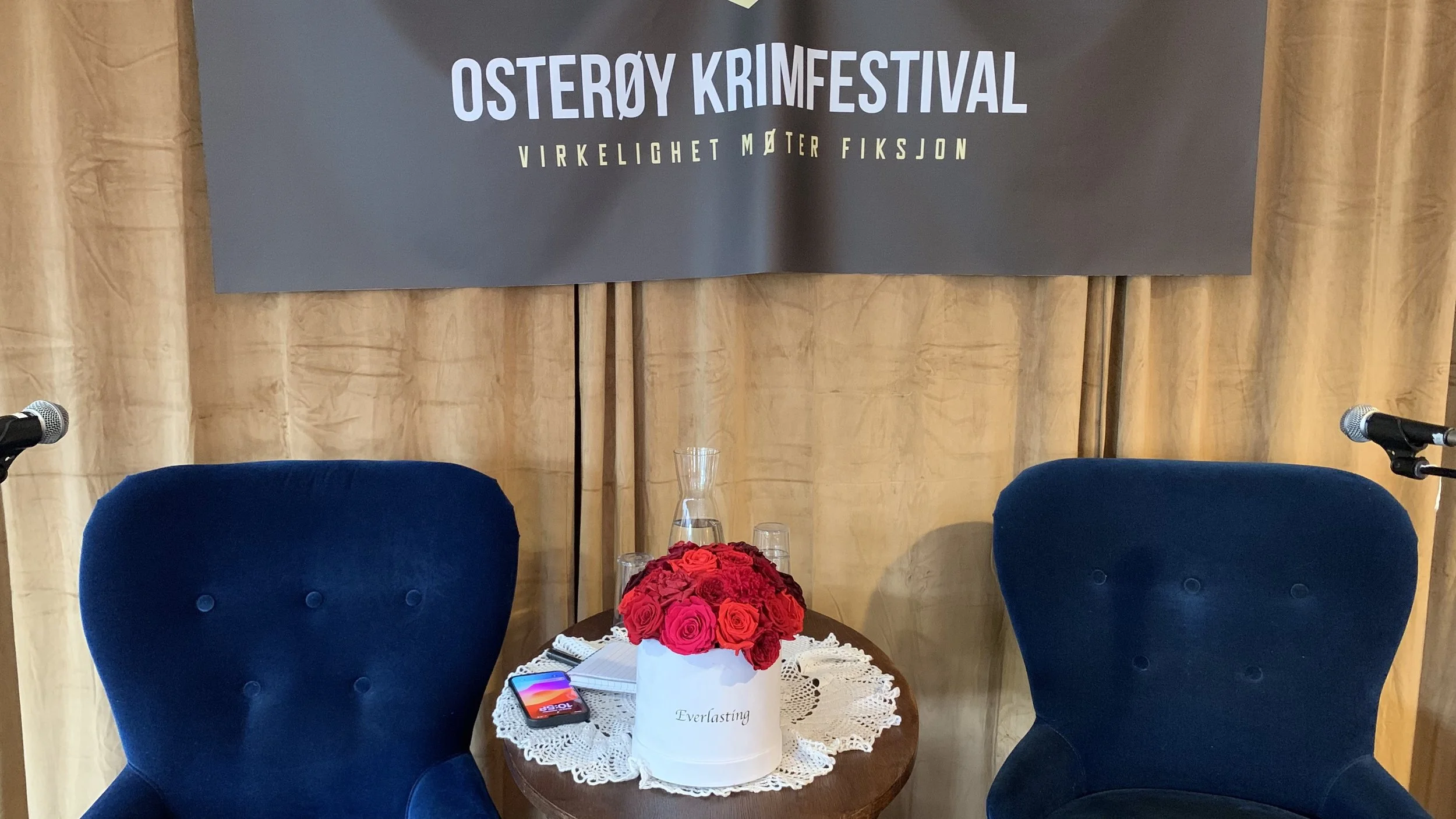 Scenebord og to blå komfortstoler foran et banner med teksten «Osterøy Krimfestival – Virkelighet møter fiksjon». På bordet står en hvit roseboks med røde roser, to glass, en vannkaraffel og et mobiltelefonetui.