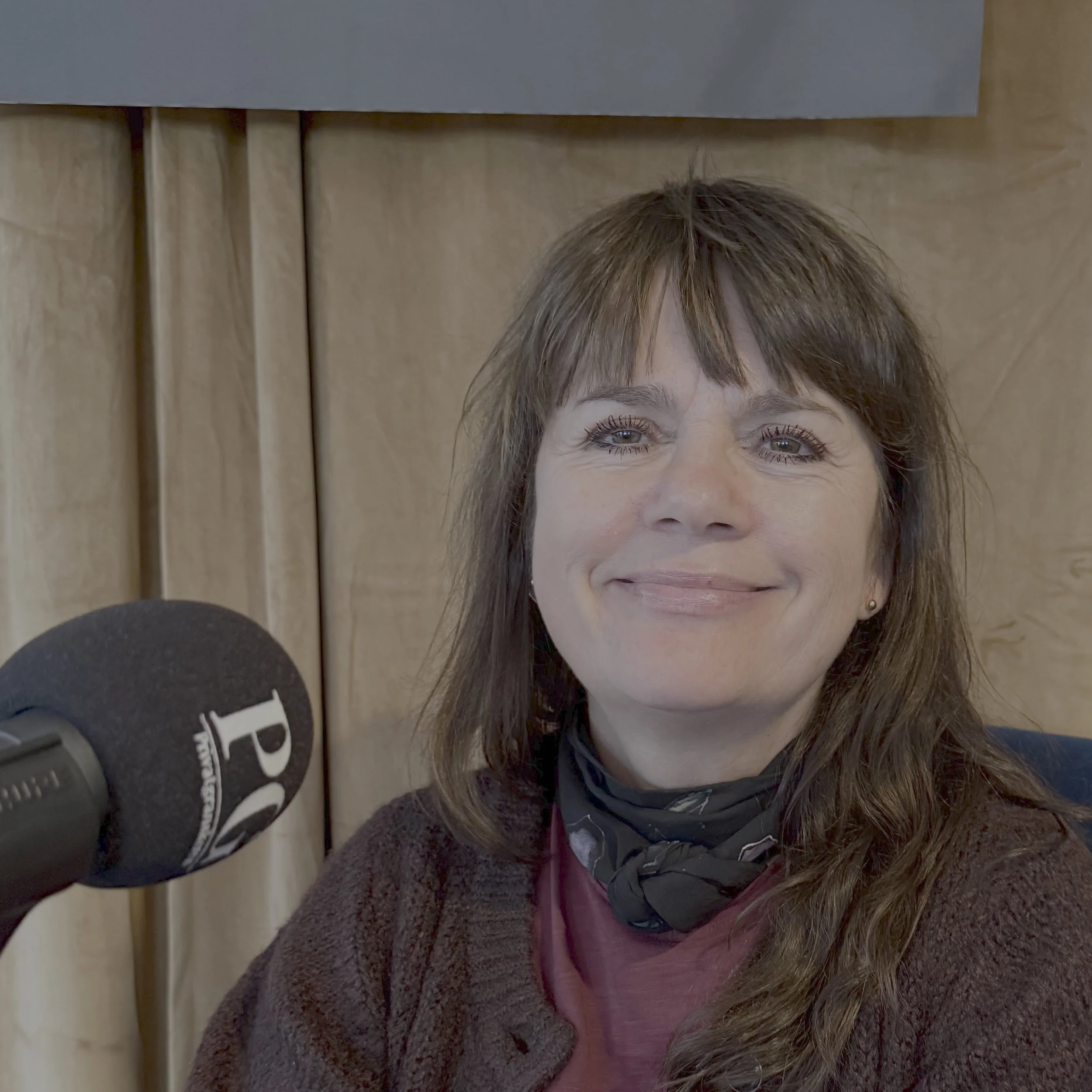 Person i podkastoppsett sitter foran en PG‑merket mikrofon, iført mørk jakke og rødfarget genser med et mørkt skjerf rundt halsen. Bakgrunnen består av lyse scenegardiner og en mørk panelplate øverst i bildet.