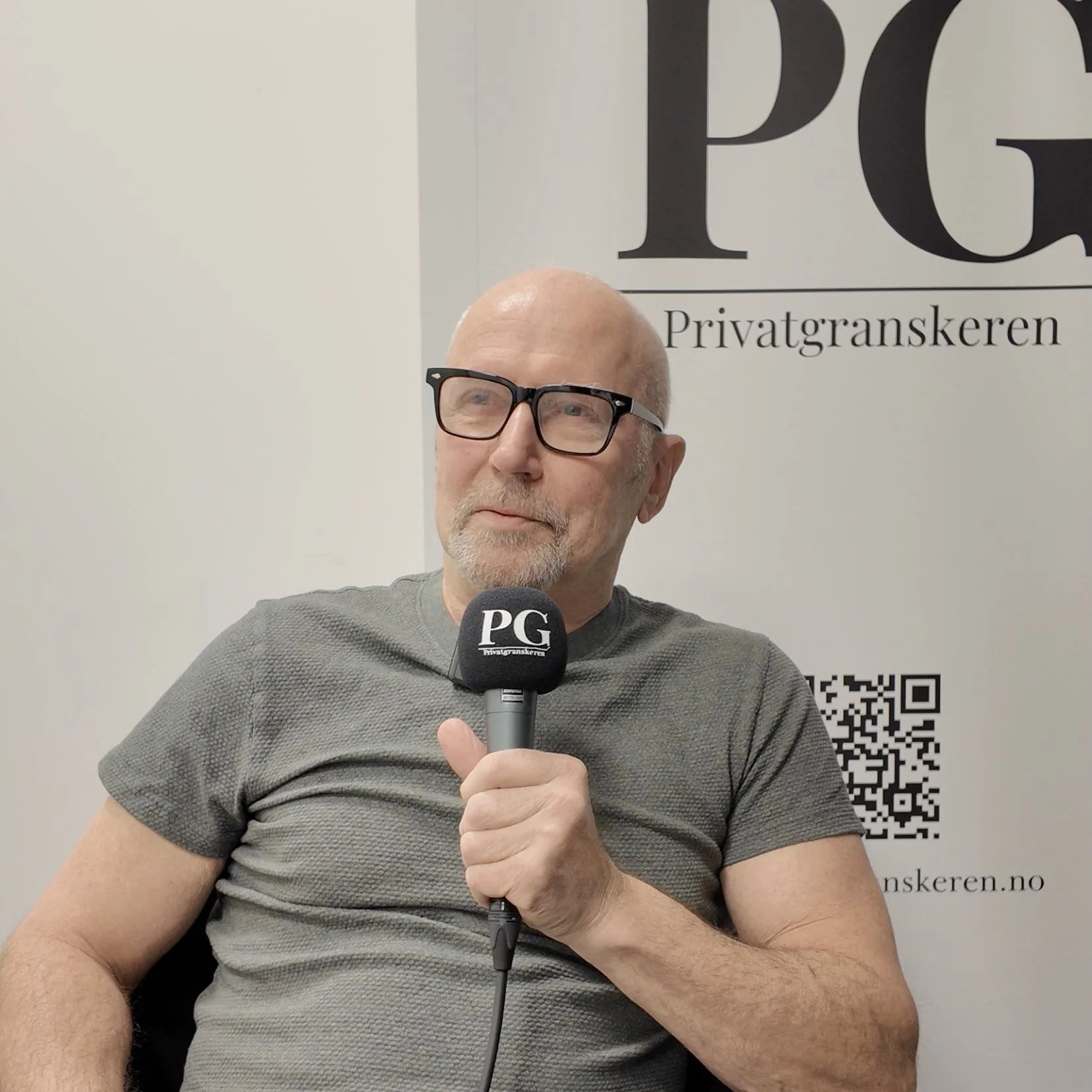 Person holder en mikrofon merket «PG» foran et banner med teksten «Privatgranskeren» og en synlig QR‑kode. Personen sitter i en stol og har på en kortermet grå genser, med lys bakgrunn.