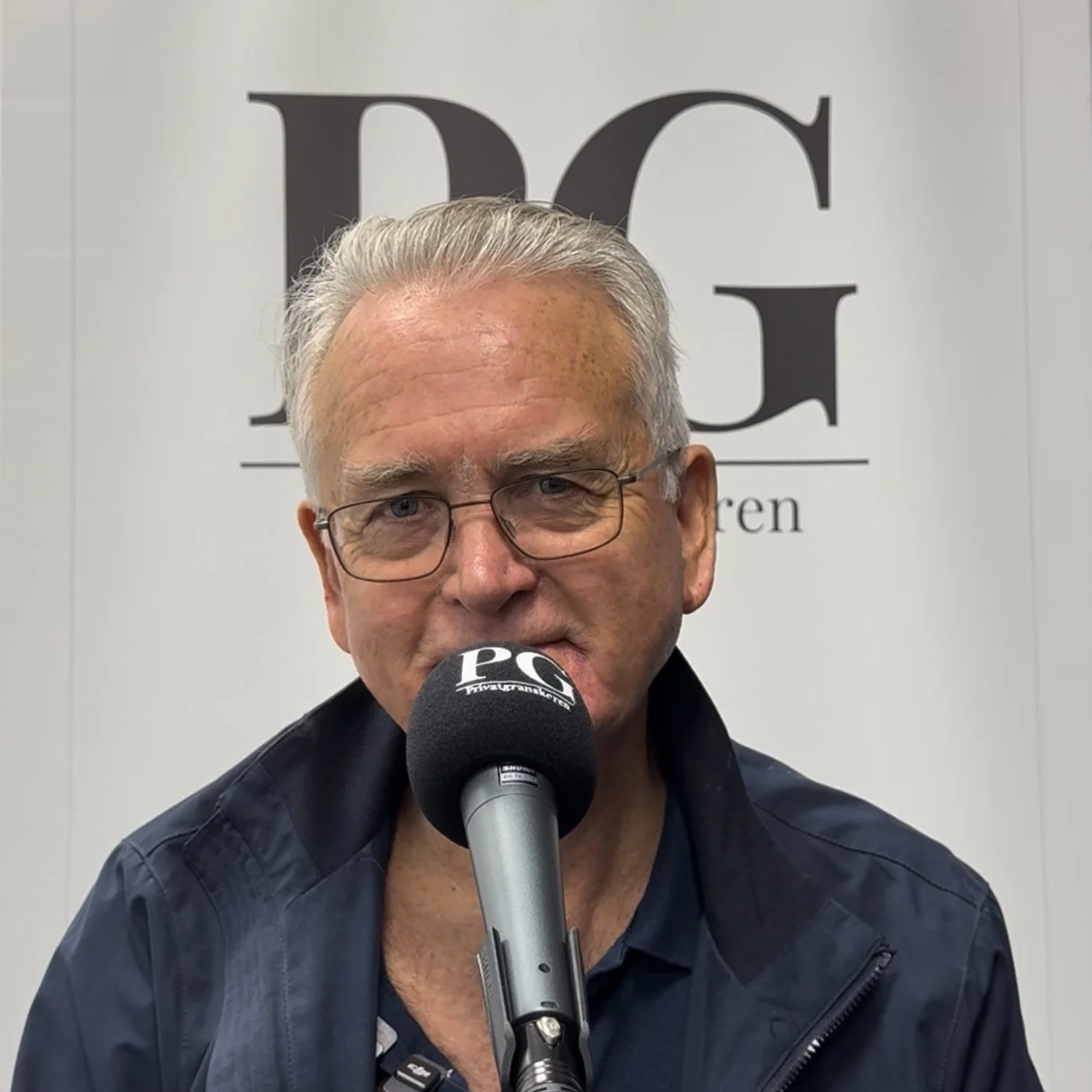 Person sitter foran en PG‑merket mikrofon i et podkastoppsett. Personen har på en mørk jakke og står foran et lyst banner med stor PG‑logo og teksten «Privatgranskeren».