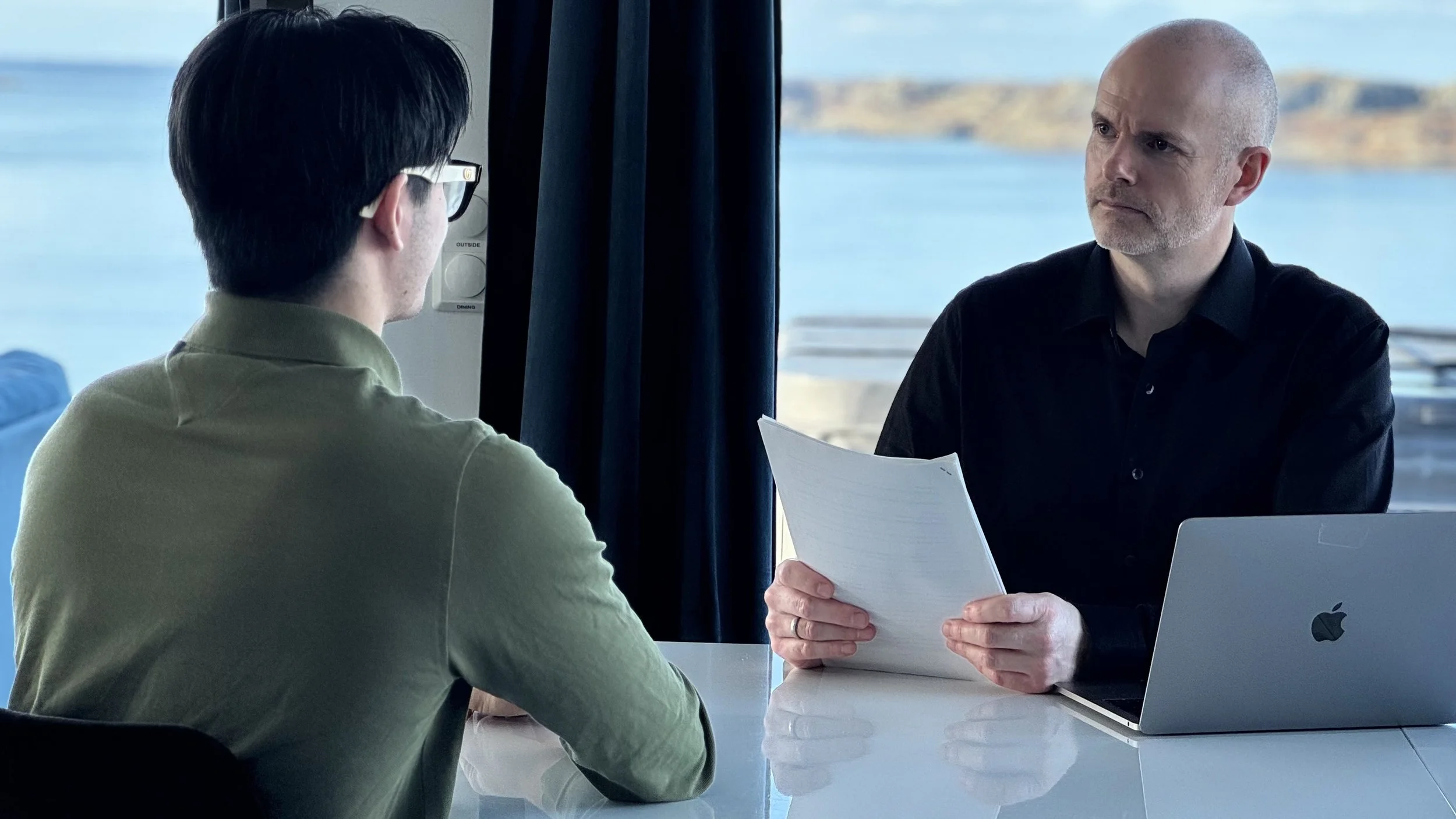 En privat etterforsker fra Privatgranskeren AS sitter ved et bord med en klient i et fysisk møte, gjennomgår dokumenter og har en laptop foran seg, med kystlandskap i bakgrunnen.