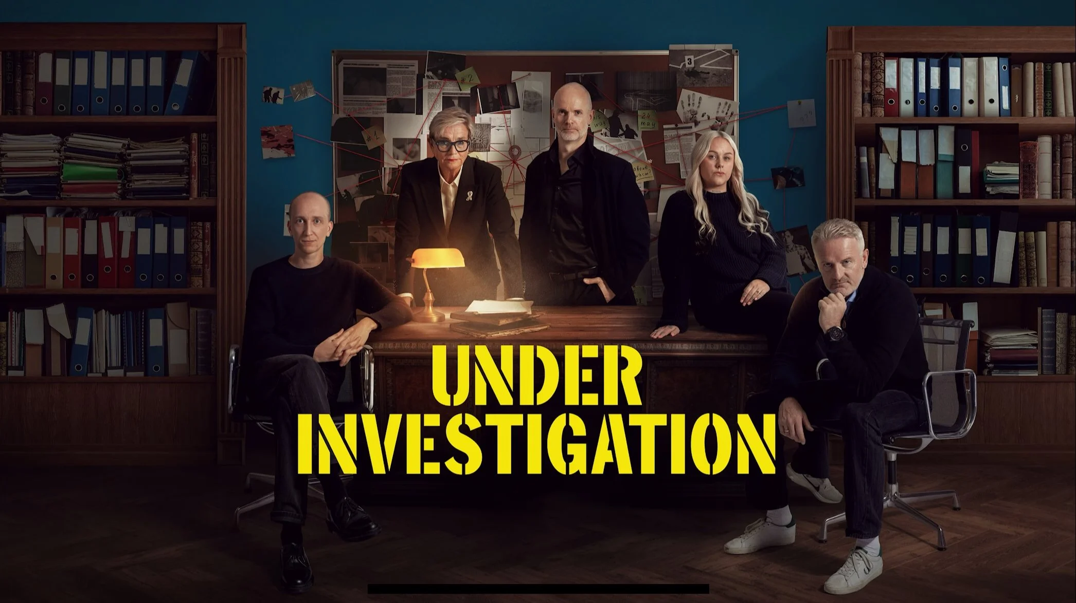 Fem personer sitter og står rundt et skrivebord i et mørkt kontorinteriør. På veggen bak henger et stort brett fylt med notater, papirer og bilder festet med tegnestifter og klyper. Rommet er omgitt av høye bokhyller fulle av permer og mapper.