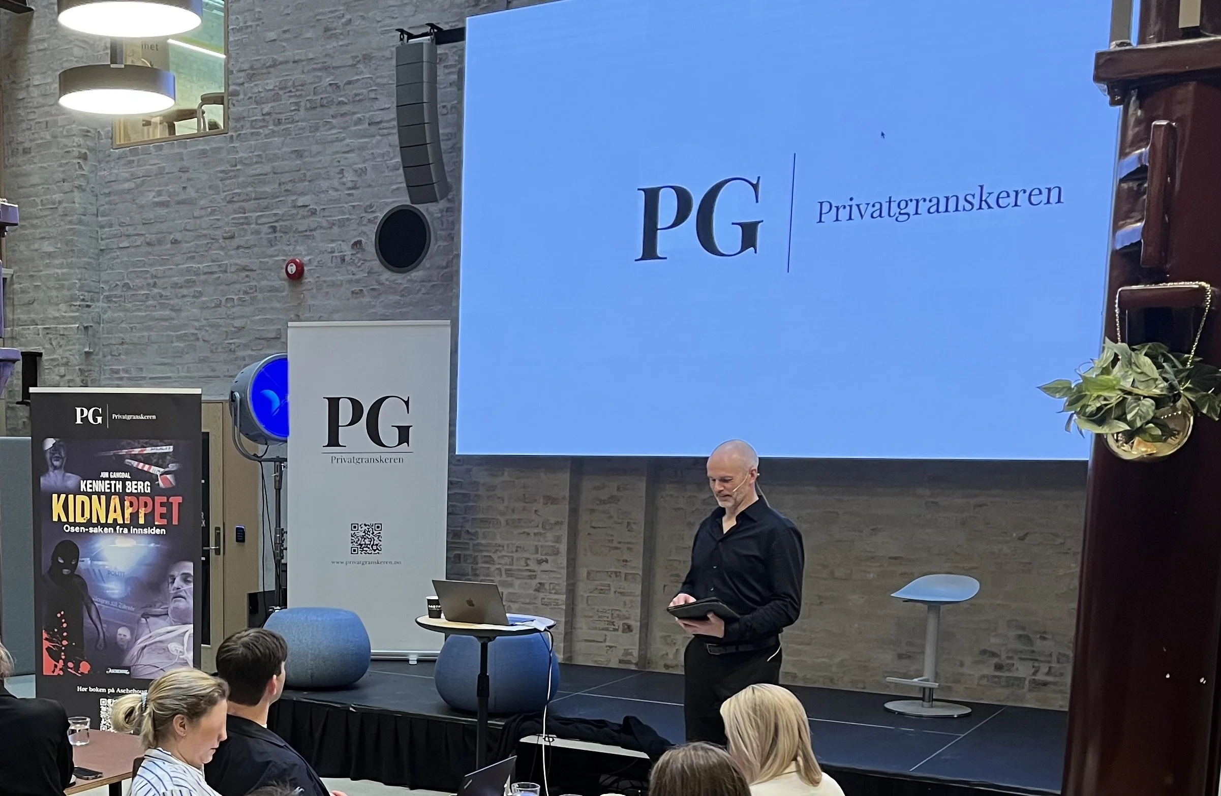 Foredragssituasjon der en person står på en scene foran en stor skjerm som viser PG logoen og teksten «Privatgranskeren». Publikum sitter ved bord foran scenen. Til venstre står bannere med PG profilering.