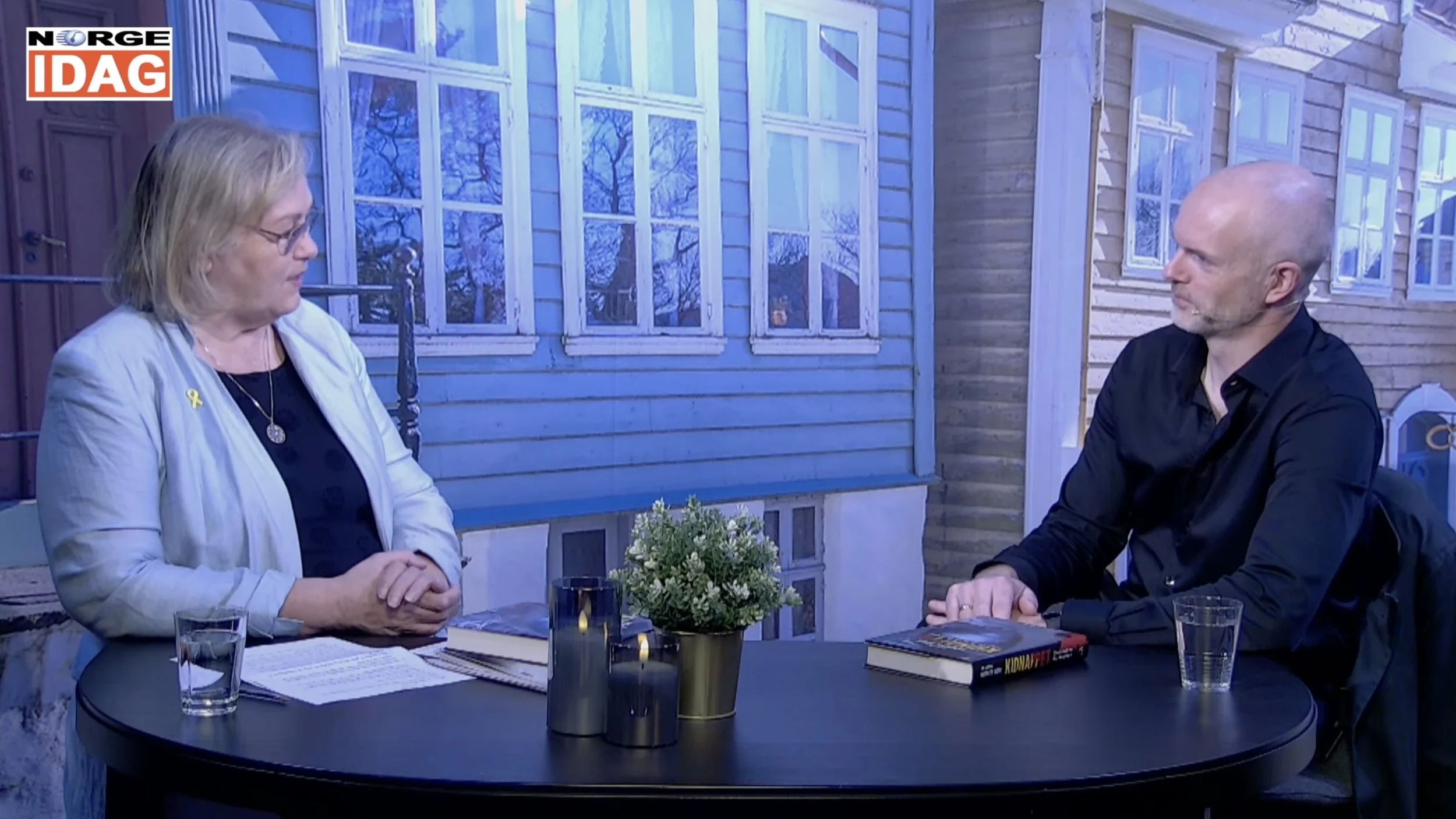 To personer sitter ved et rundt bord i et studio under et intervju for Norge IDAG. På bordet ligger en bok, flere glass med vann og dekorlys, samt en liten grønn plante. Bak dem vises en bakgrunn som forestiller fasaden av trehus med vinduer.