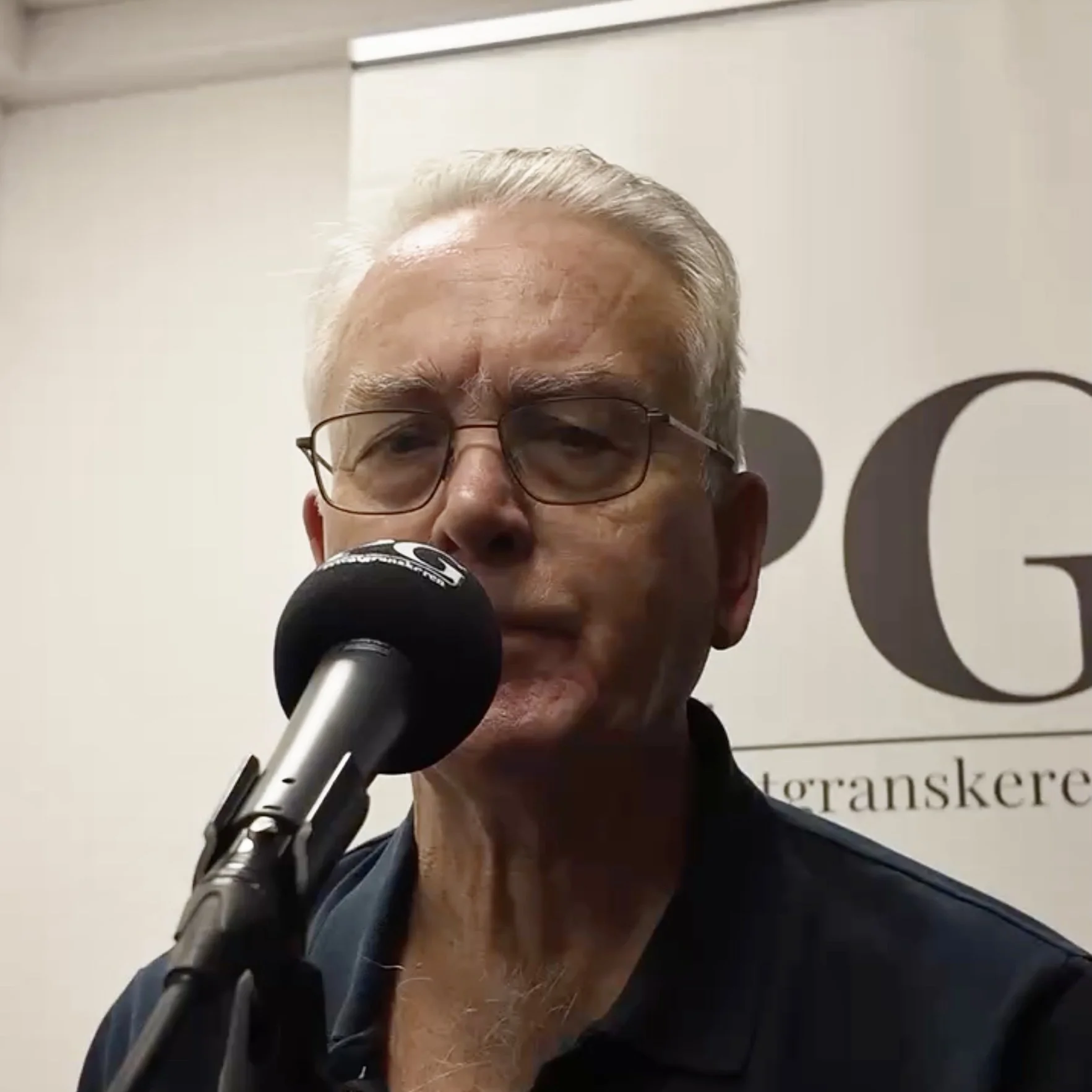 Person i podkaststudio med store hodetelefoner og mikrofon plassert foran seg. Bakgrunnen viser et lyst banner med store bokstaver fra logoen til Privatgranskeren.