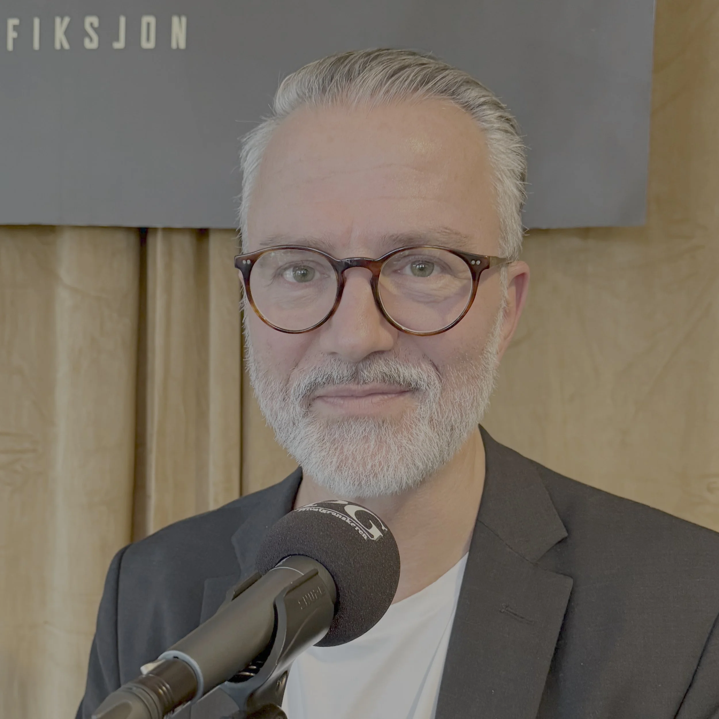 Person i et podkaststudio sitter foran en PG‑merket mikrofon, iført en mørk blazer og lys T‑skjorte. Bakgrunnen består av et lyst sceneteppe og en mørkere panelplate med delvis synlig tekst.