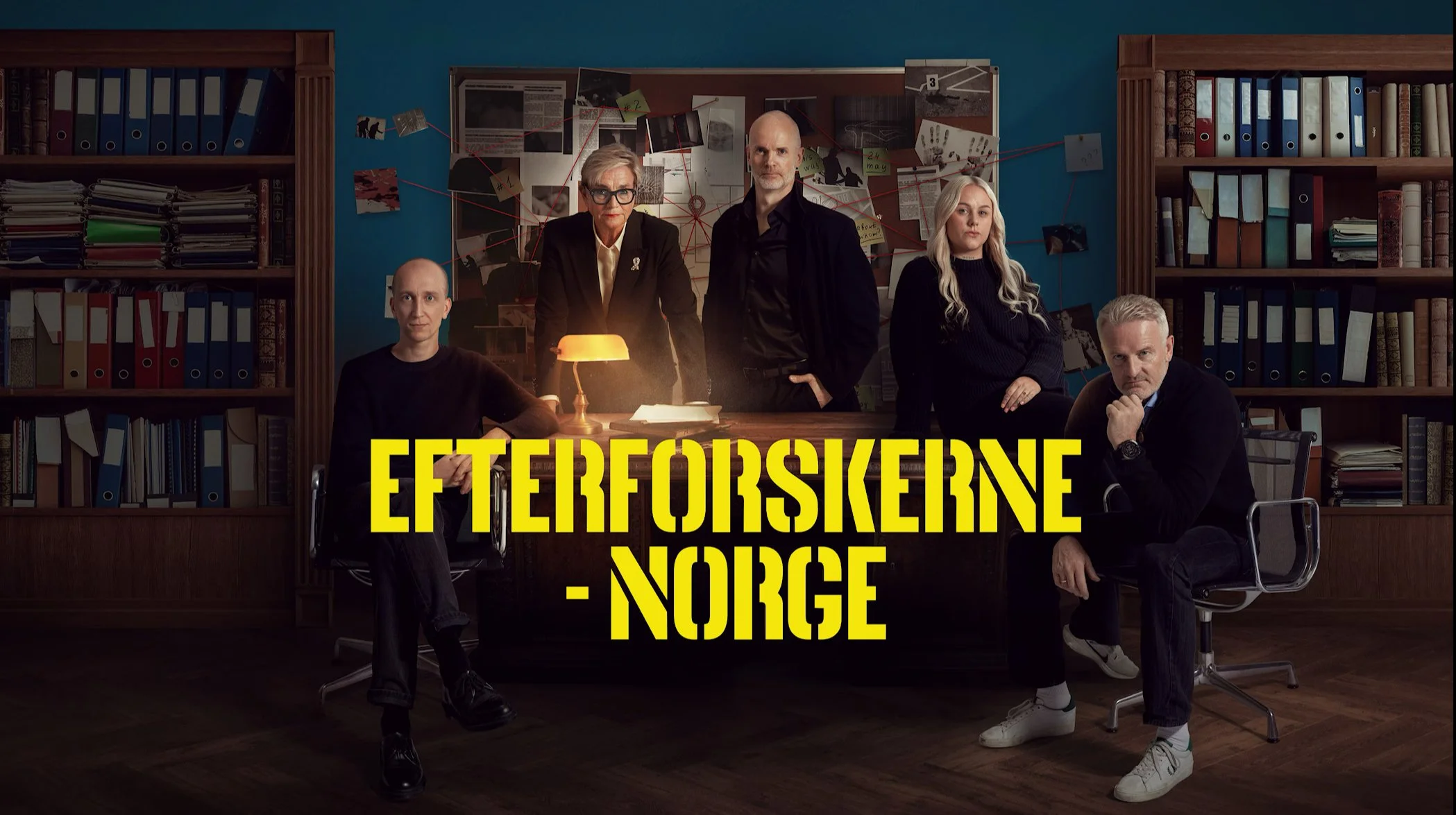 Fem personer sitter og står rundt et skrivebord i et mørkt kontorinteriør. På veggen bak henger et stort brett fylt med notater, papirer og bilder festet med tegnestifter og klyper. Rommet er omgitt av høye bokhyller fulle av permer og mapper.