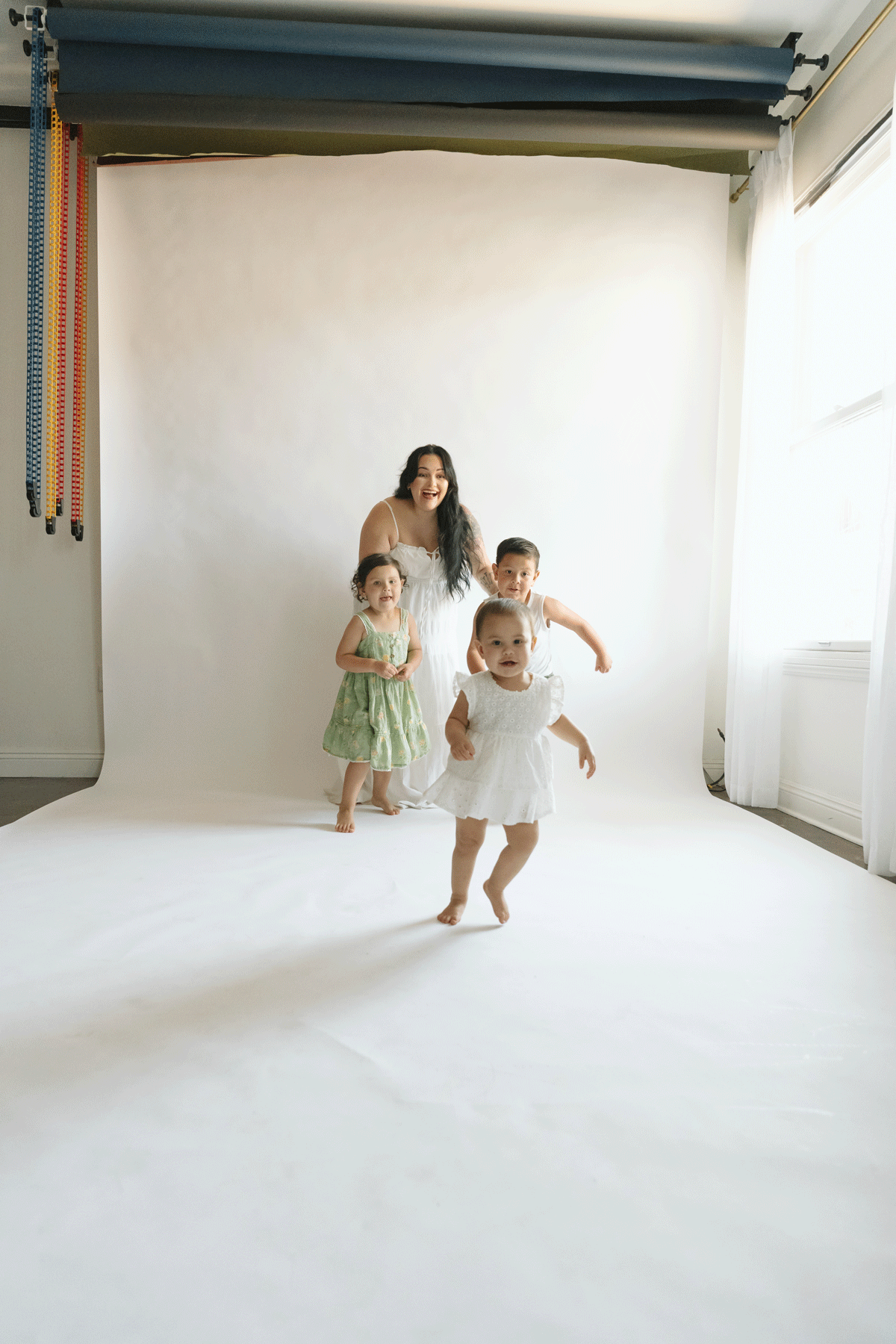 Woman with three young children in a photography studio, white backdrop, colorful rolls overhead.