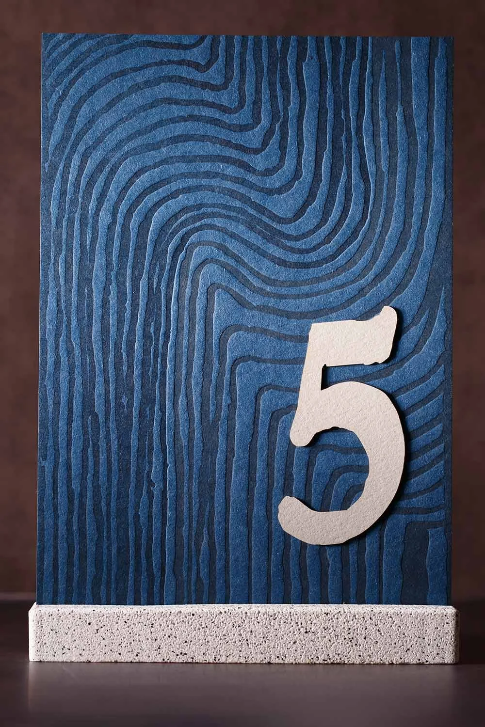 Custom wedding table number featuring layered blue paper, letterpress-inspired topographic texture, dimensional numeral, and a stone-inspired display base.
