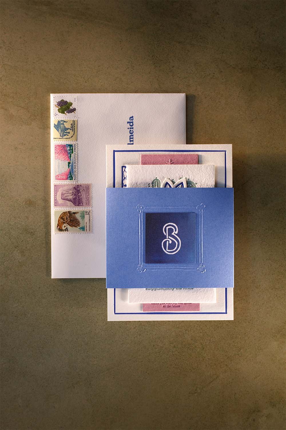 Custom blue wedding invitation suite with embossed monogram belly band, vintage stamps, and textured cotton paper details.