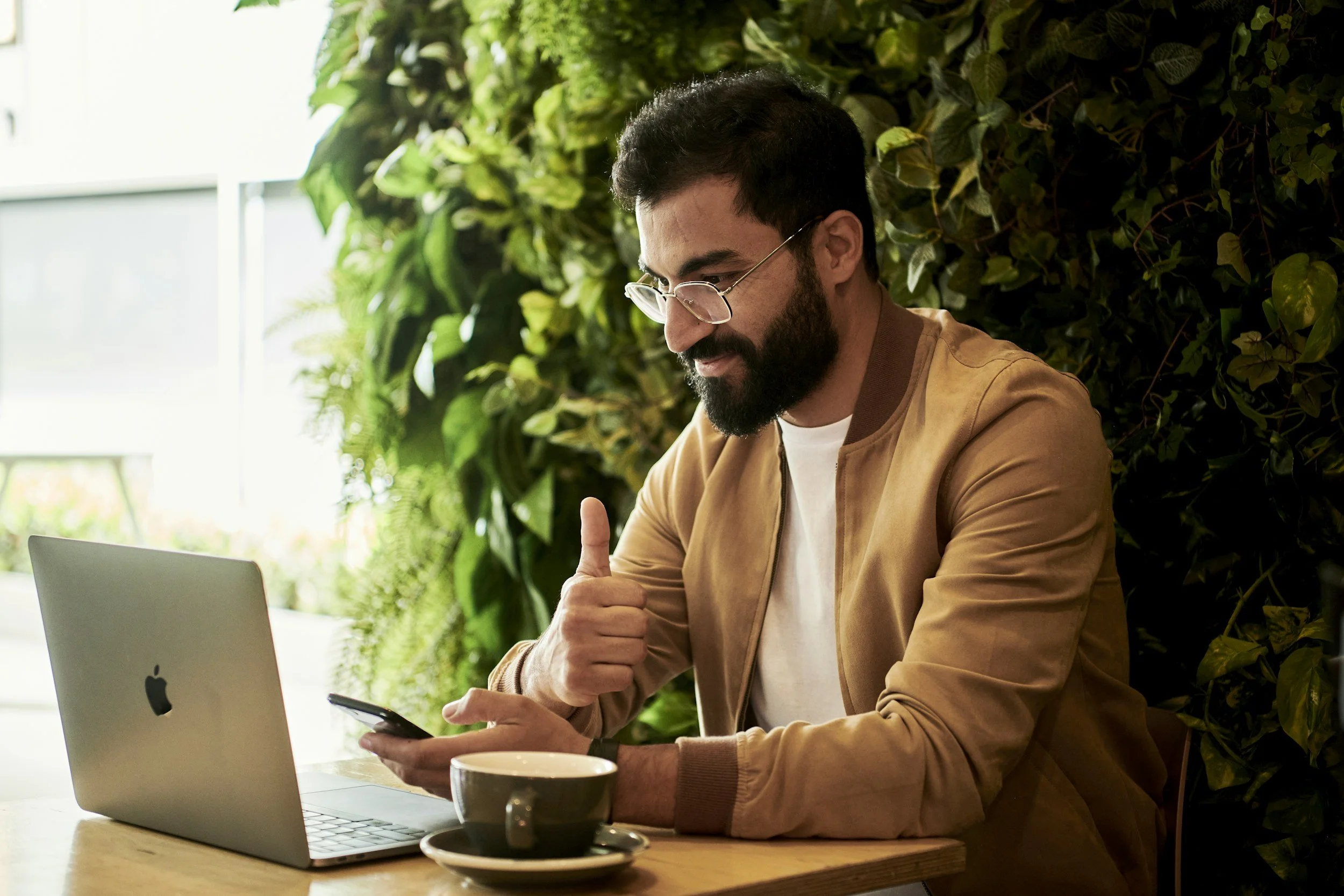 A man with glasses and a beard sitting at a table in a café, giving a thumbs-up while using his smartphone, with a MacBook laptop and a coffee cup on the table, and a green plant wall in the background.