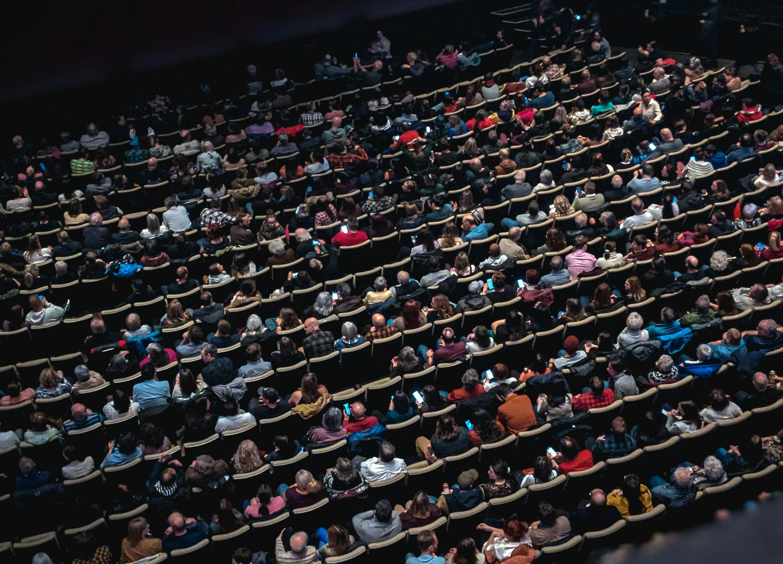 An audience sitting in theater seats, many using their phones.