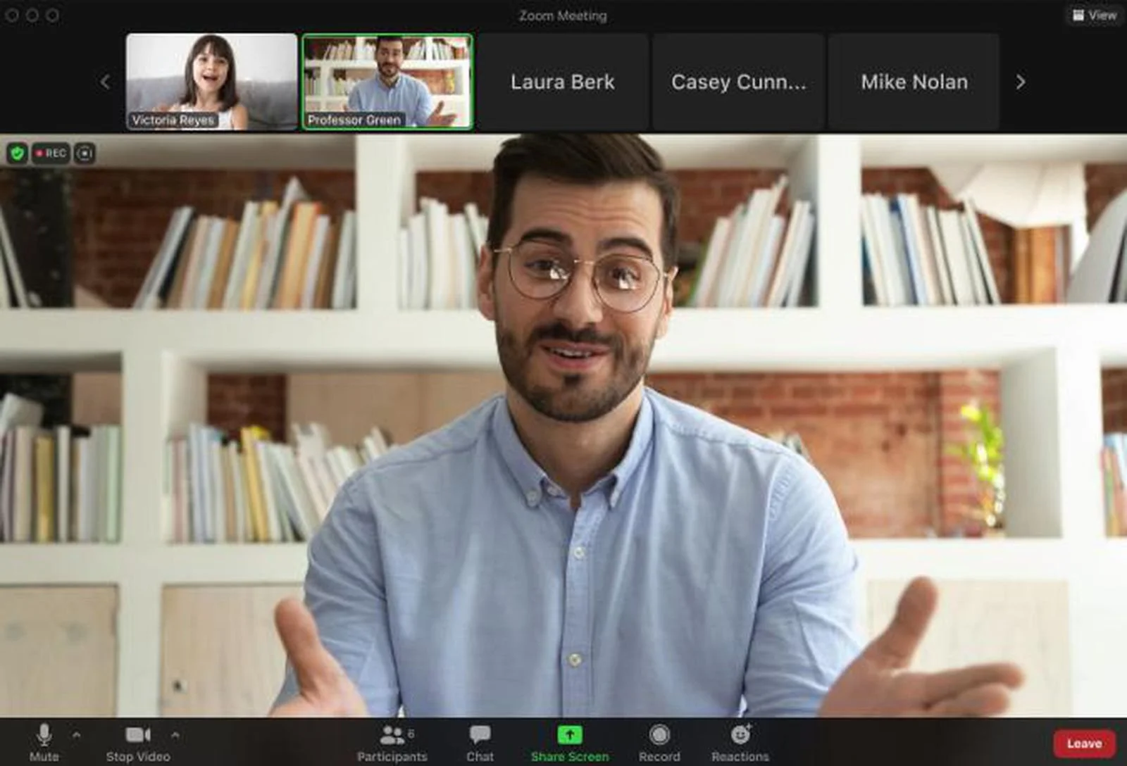 A man with glasses and a beard, wearing a light blue shirt, is speaking during a Zoom meeting. The background features white shelving with books and a brick wall.