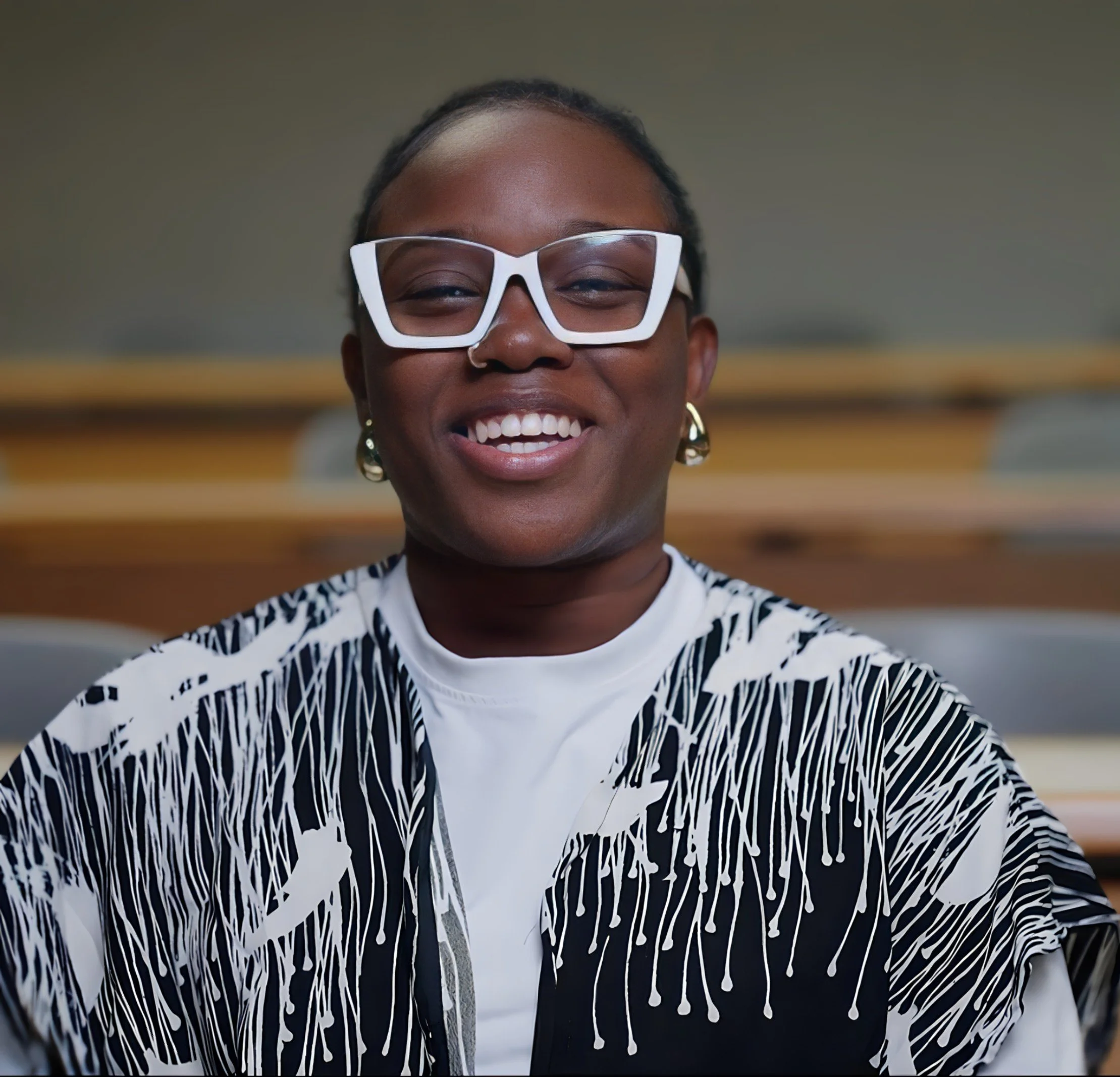Close-up of a smiling woman wearing white glasses, gold earrings, and a black and white patterned jacket in an indoor setting.