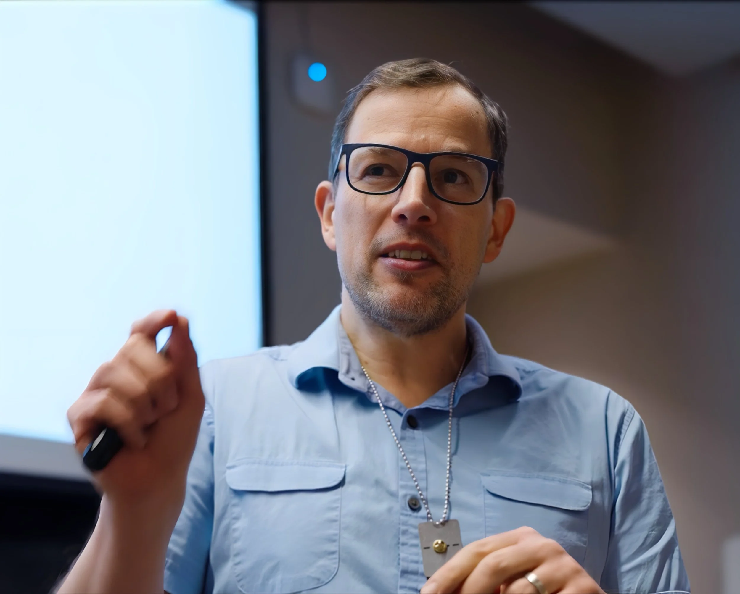 A man wearing glasses and a light blue shirt, holding a black marker, speaking in front of a presentation screen.