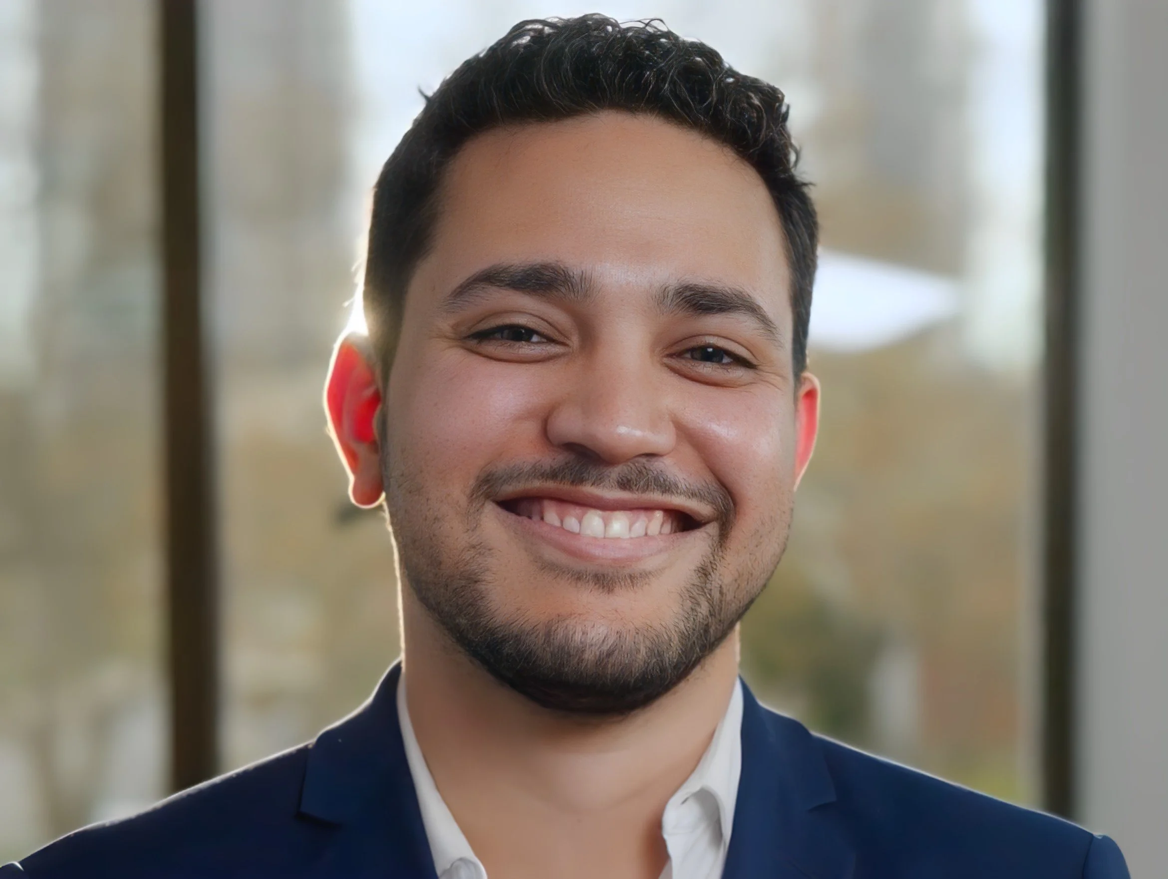 A smiling man in a suit with a blurred window background.