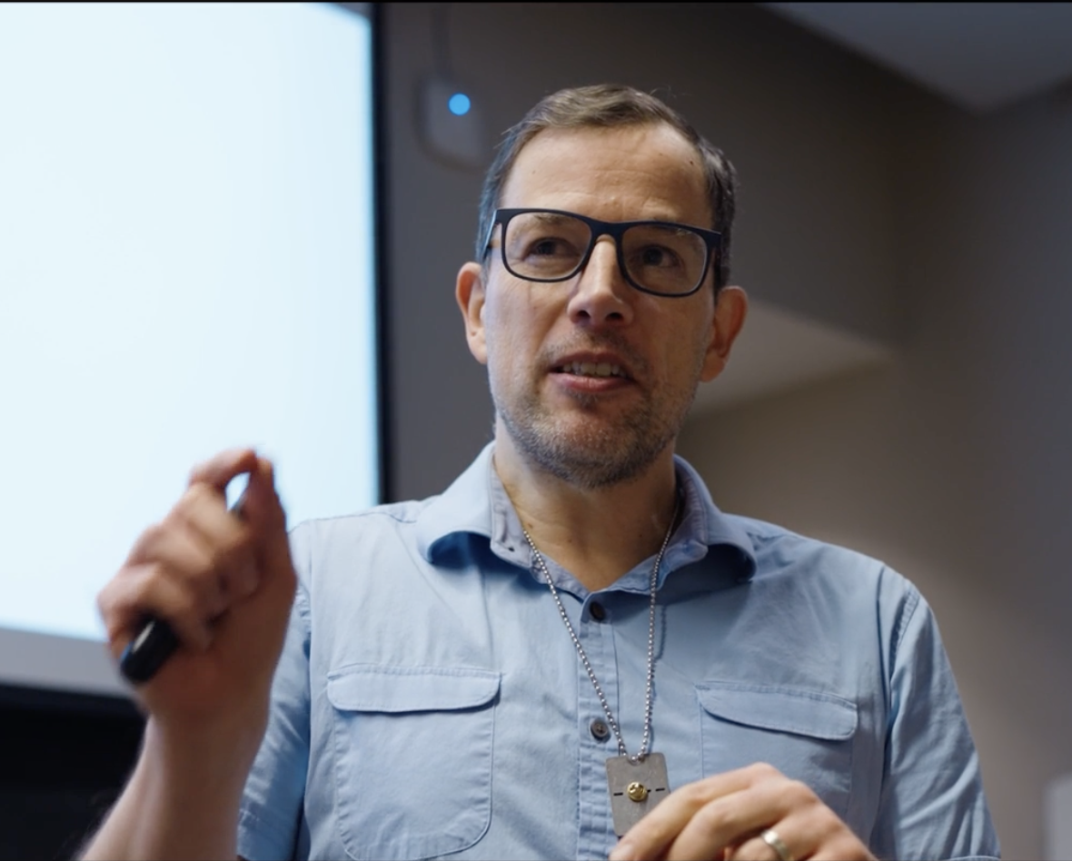 A middle-aged man with glasses and a beard wearing a light blue shirt, speaking and gesturing with his left hand, holding a small object in his right hand, in a presentation or classroom setting.