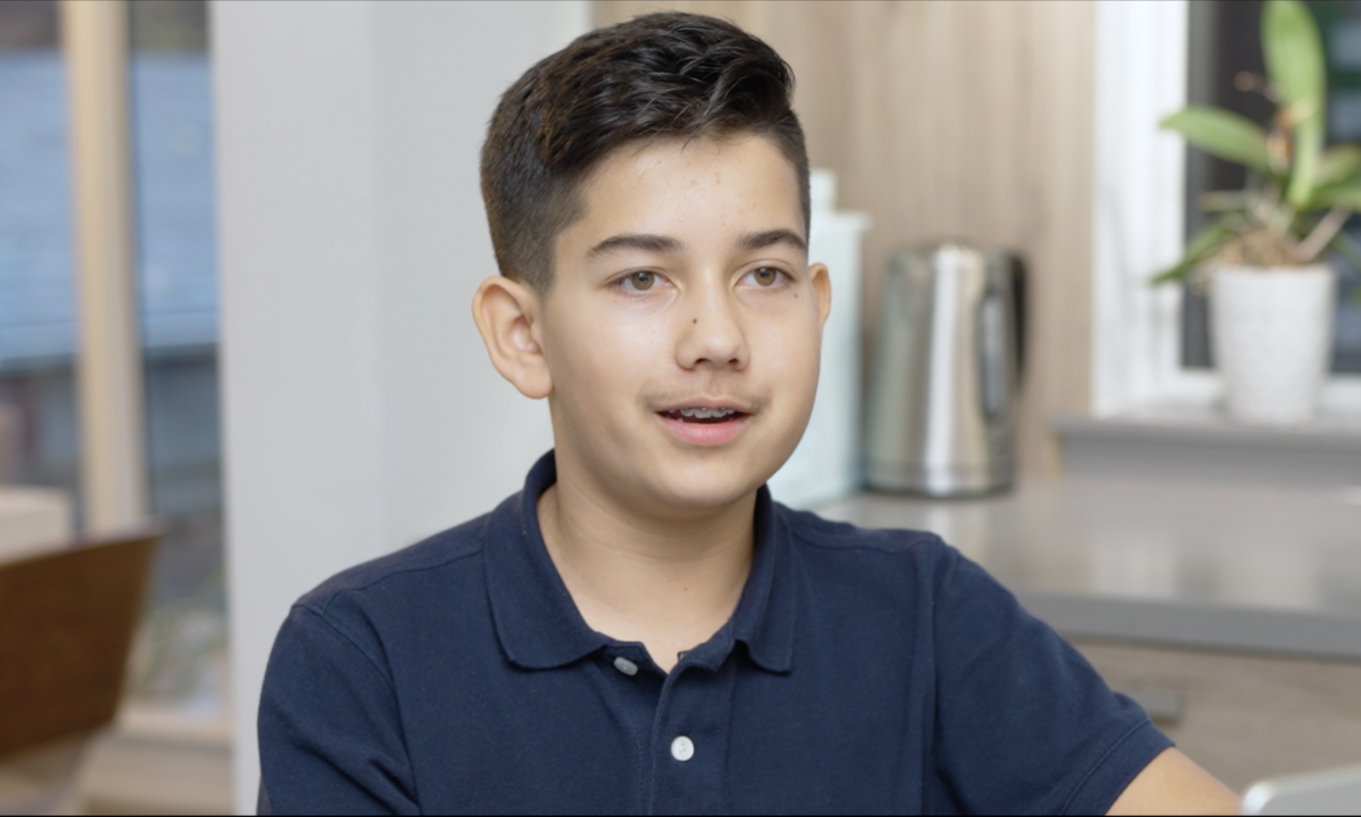 A young boy with dark hair, wearing a blue polo shirt, sitting indoors with a neutral expression. In the background, there is a potted plant and a countertop with a metallic kettle.