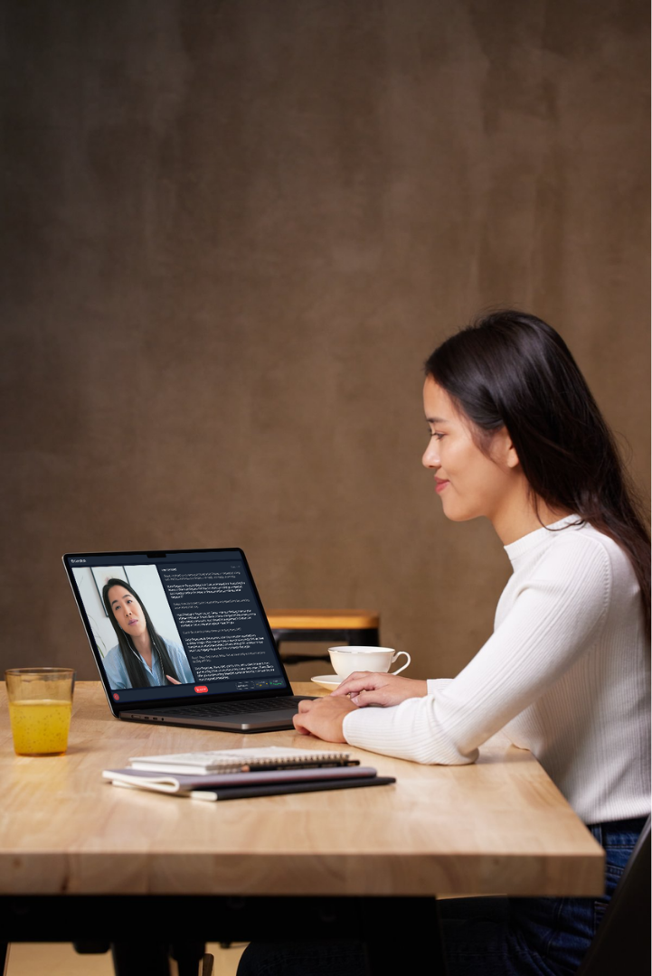 Woman in white sweater having a video call on laptop at a wooden table with notebooks, a cup of coffee, and a glass of orange juice.