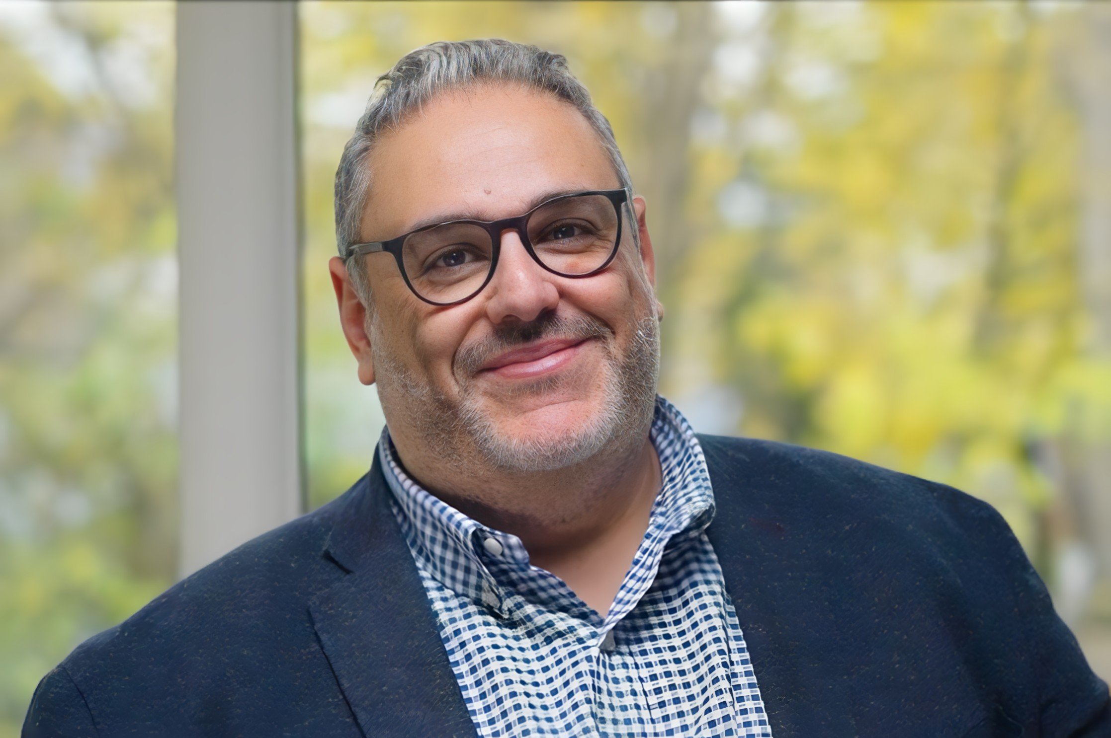 A smiling middle-aged man with gray hair and beard, wearing glasses, a dark blazer, and a checkered shirt, standing in front of a window with trees outside.