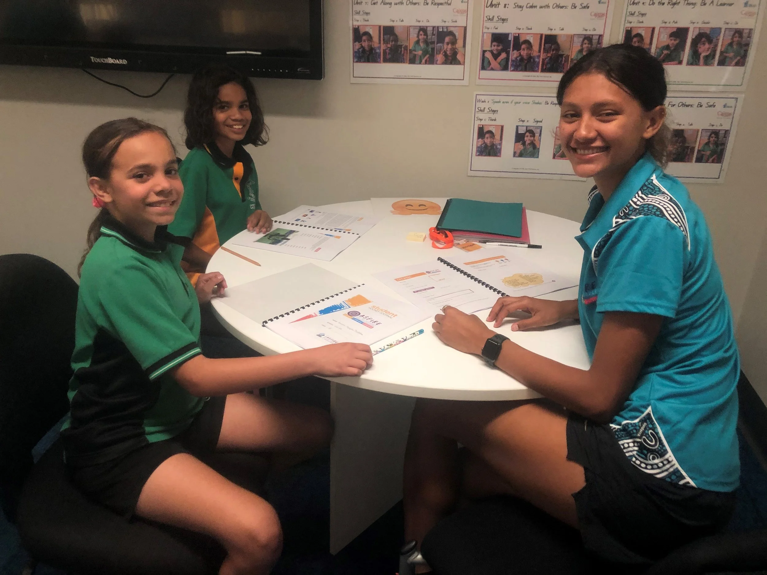 Three girls sitting around a white table in a classroom, smiling at the camera. They are examining educational booklets and papers. The classroom has posters on the wall and a large monitor.