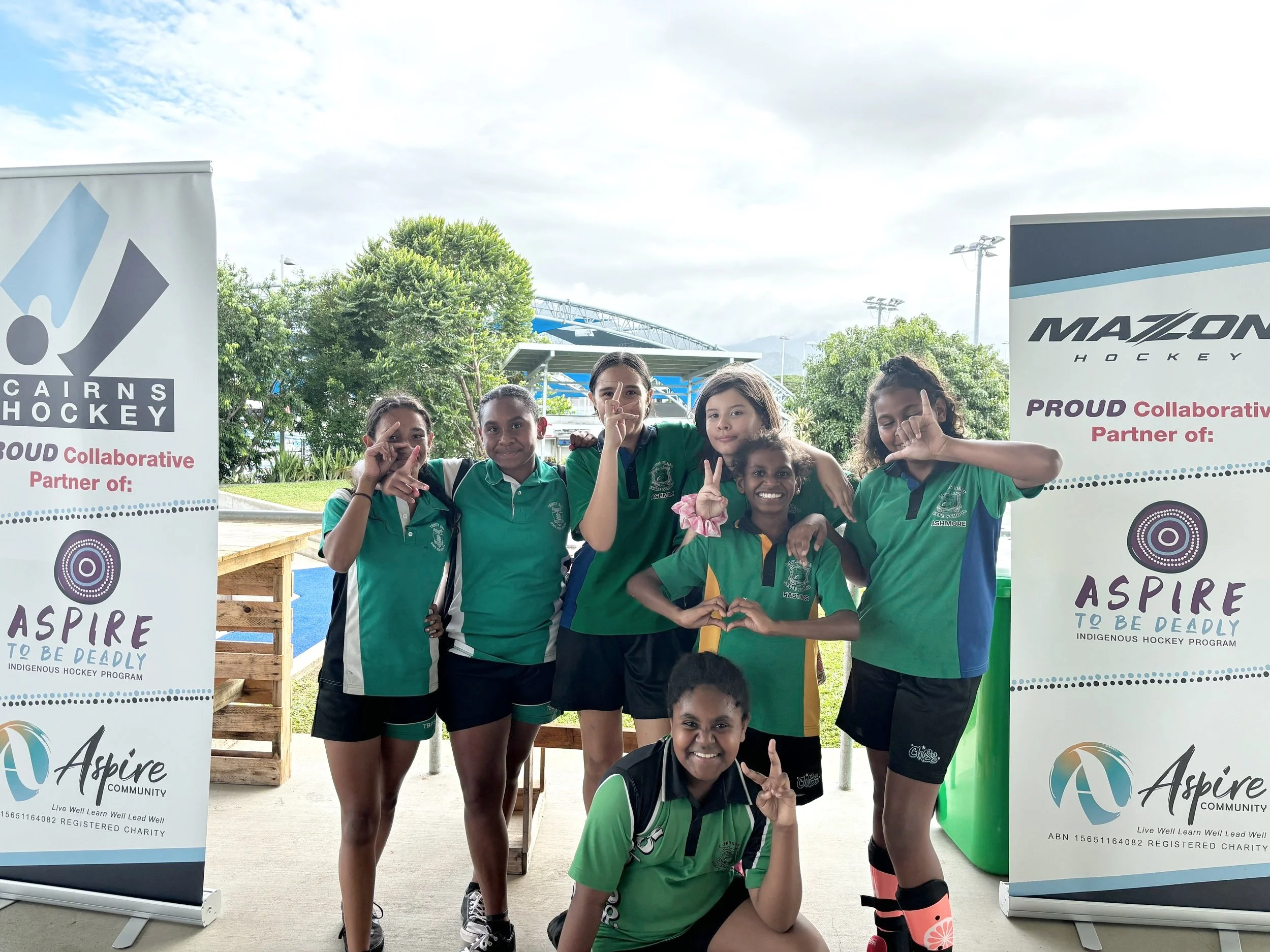 A group of seven kids in green and blue hockey uniforms posing outdoors, making hand gestures, with two banners promoting Indigenous hockey collaboration and community program, in a park with trees and a cloudy sky.