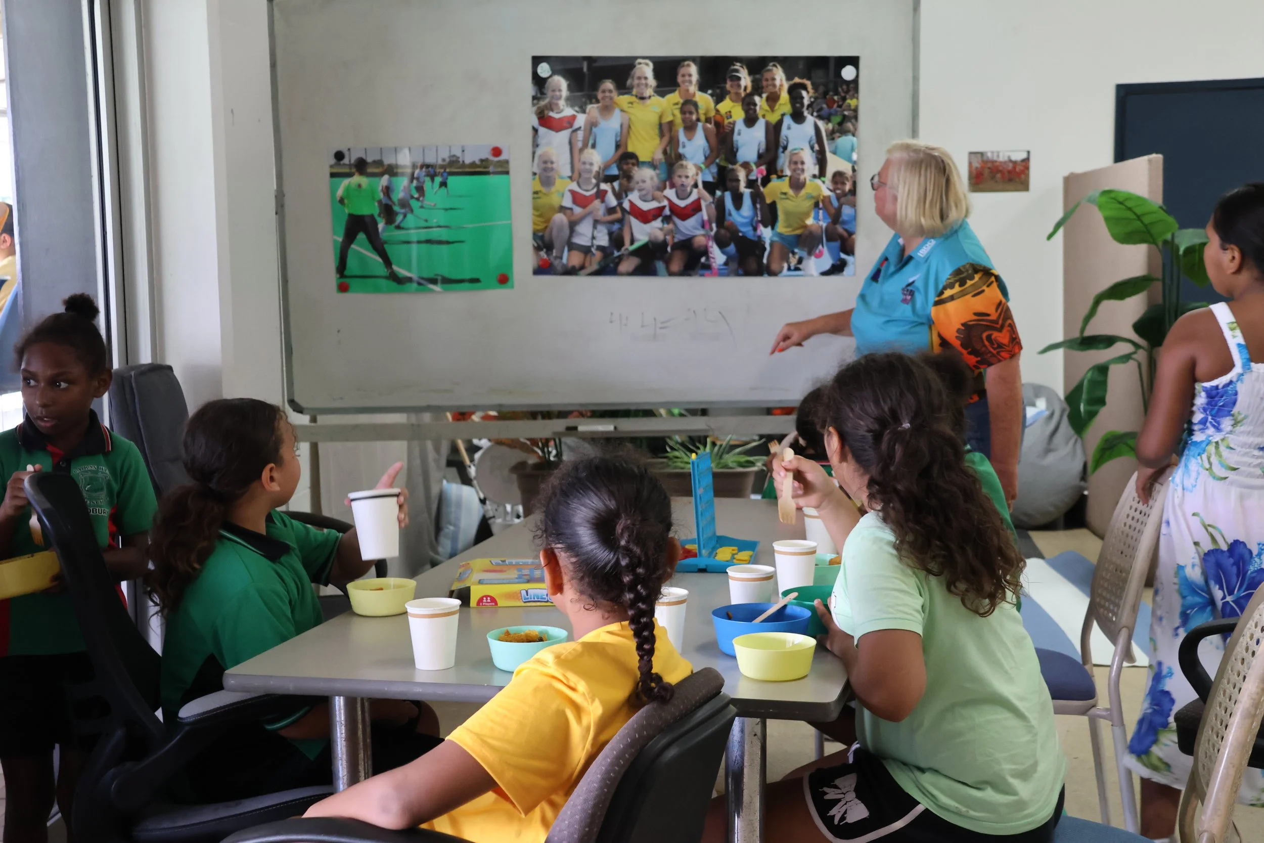 A classroom scene with children sitting around a table, a teacher pointing to photos on a whiteboard, with posters and plants in the background.