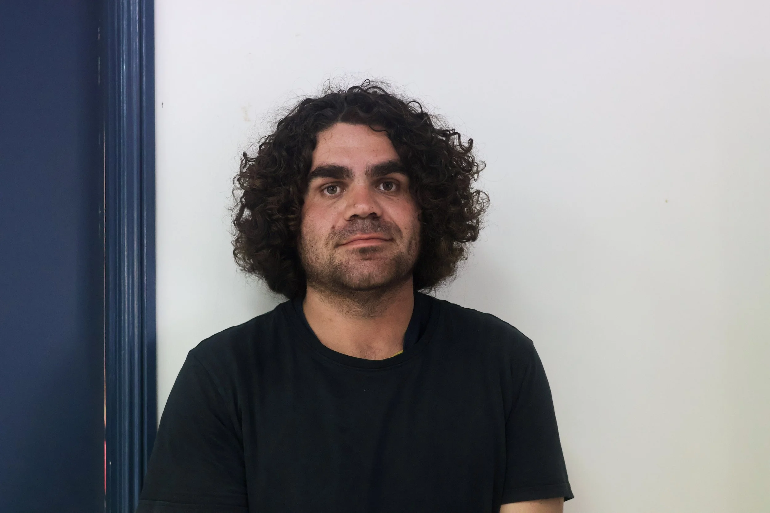 A man with curly dark hair and light skin, wearing a black T-shirt, sitting in front of a white wall with a blue door frame on the left.