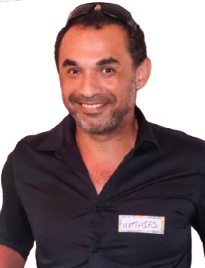 A man with short dark hair and a beard, smiling, wearing a black collared shirt with a name tag standing against a plain background.