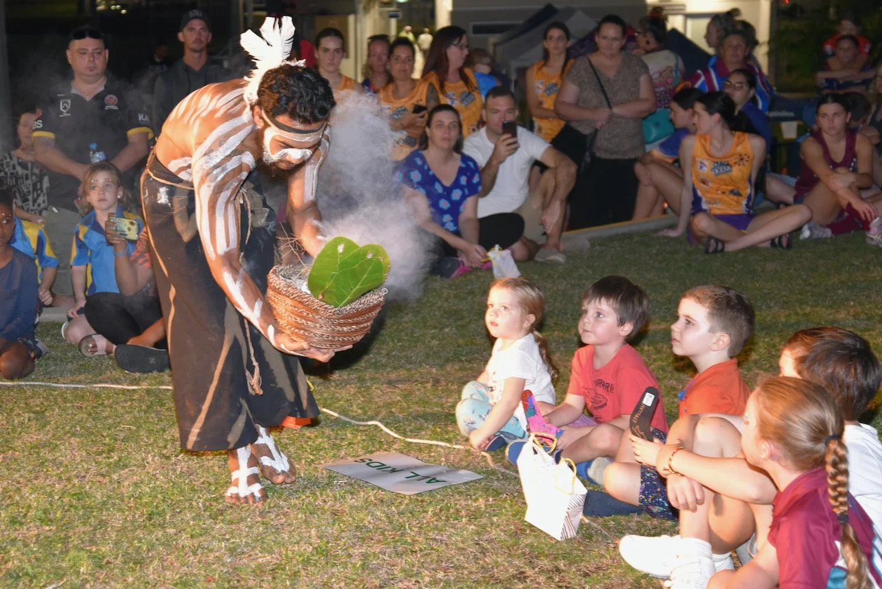 A performer dressed in traditional indigenous attire with face paint, holding a basket with a large green leaf, is performing a ritual or dance in front of an audience of children and adults sitting and watching outdoors during nighttime.
