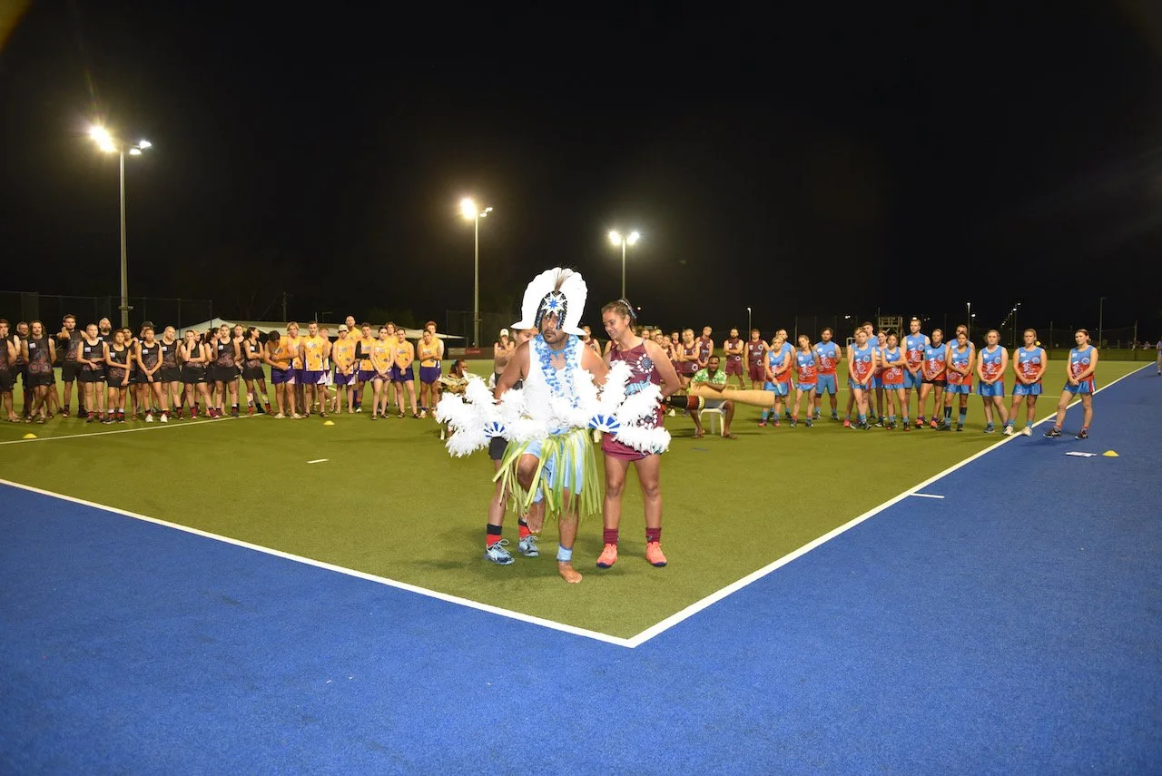 A group of women in sports uniforms standing on a sports field at night, with a person in elaborate costume in the foreground.