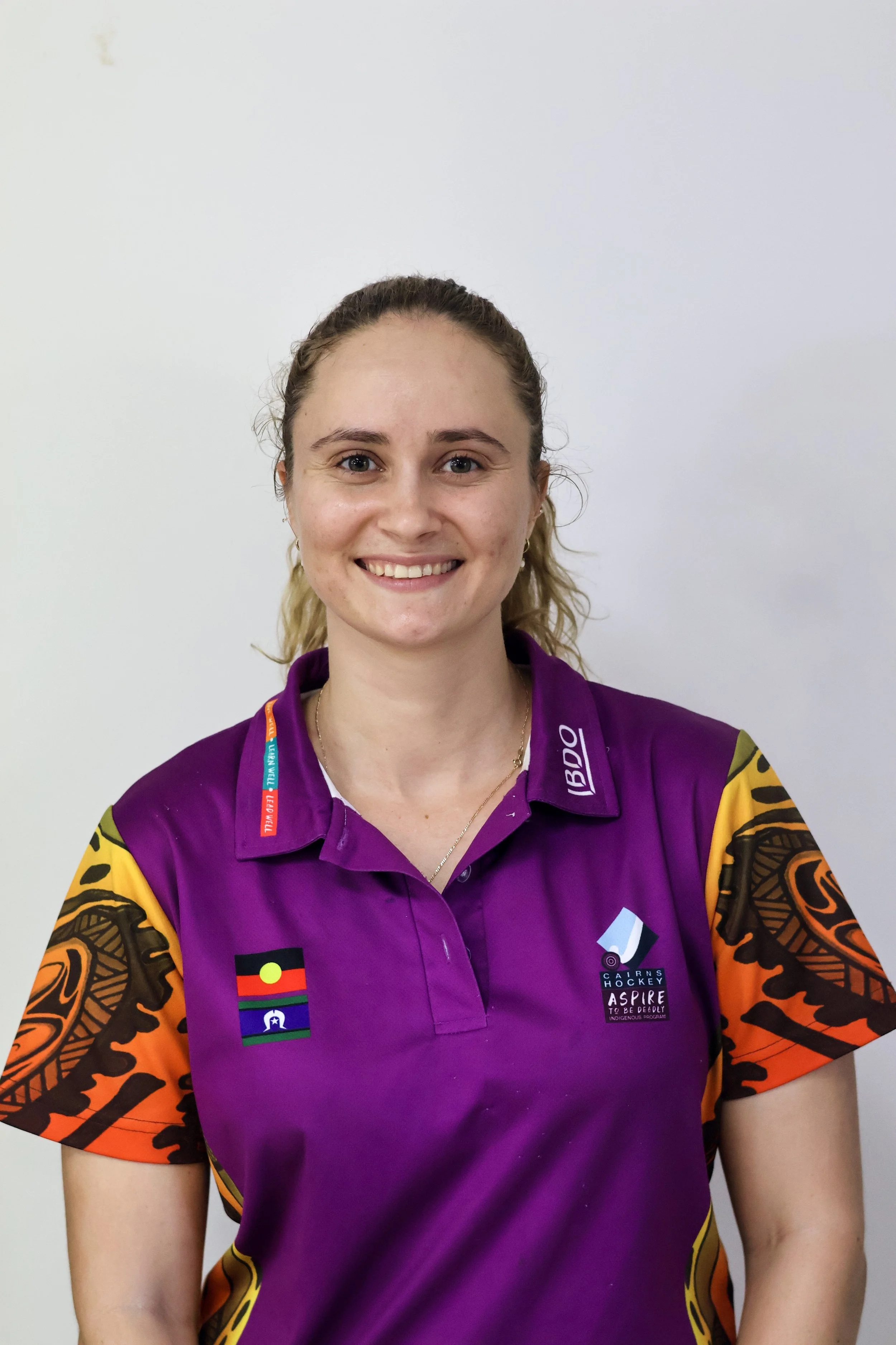 A young woman with blonde hair in a ponytail, smiling, wearing a purple Aboriginal-themed sports jersey with Indigenous flags and logos, standing against a plain white background.