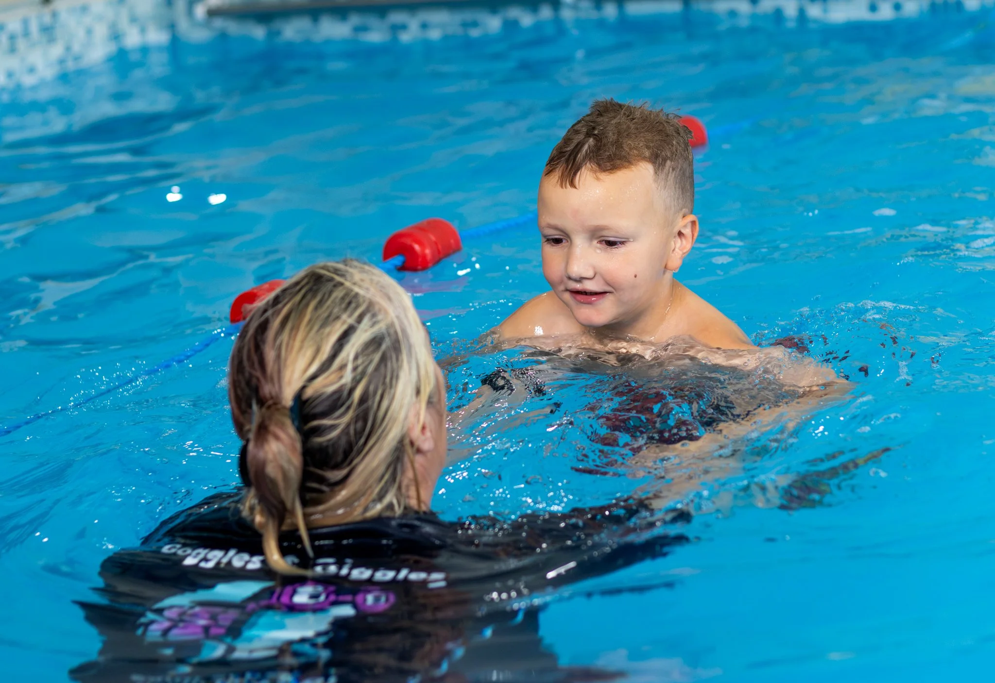 A young boy in a swimming lesson with a swim teacher, with red and blue floating lane dividers in the background.