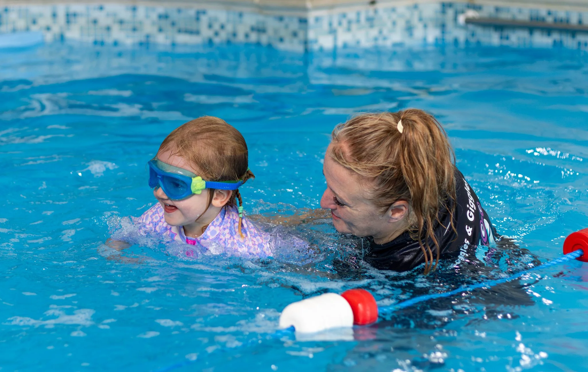 A young girl with blonde hair wearing blue swimming goggles learning to swim with an instructor in a blue tiled indoor pool, holding onto a pool noodle.