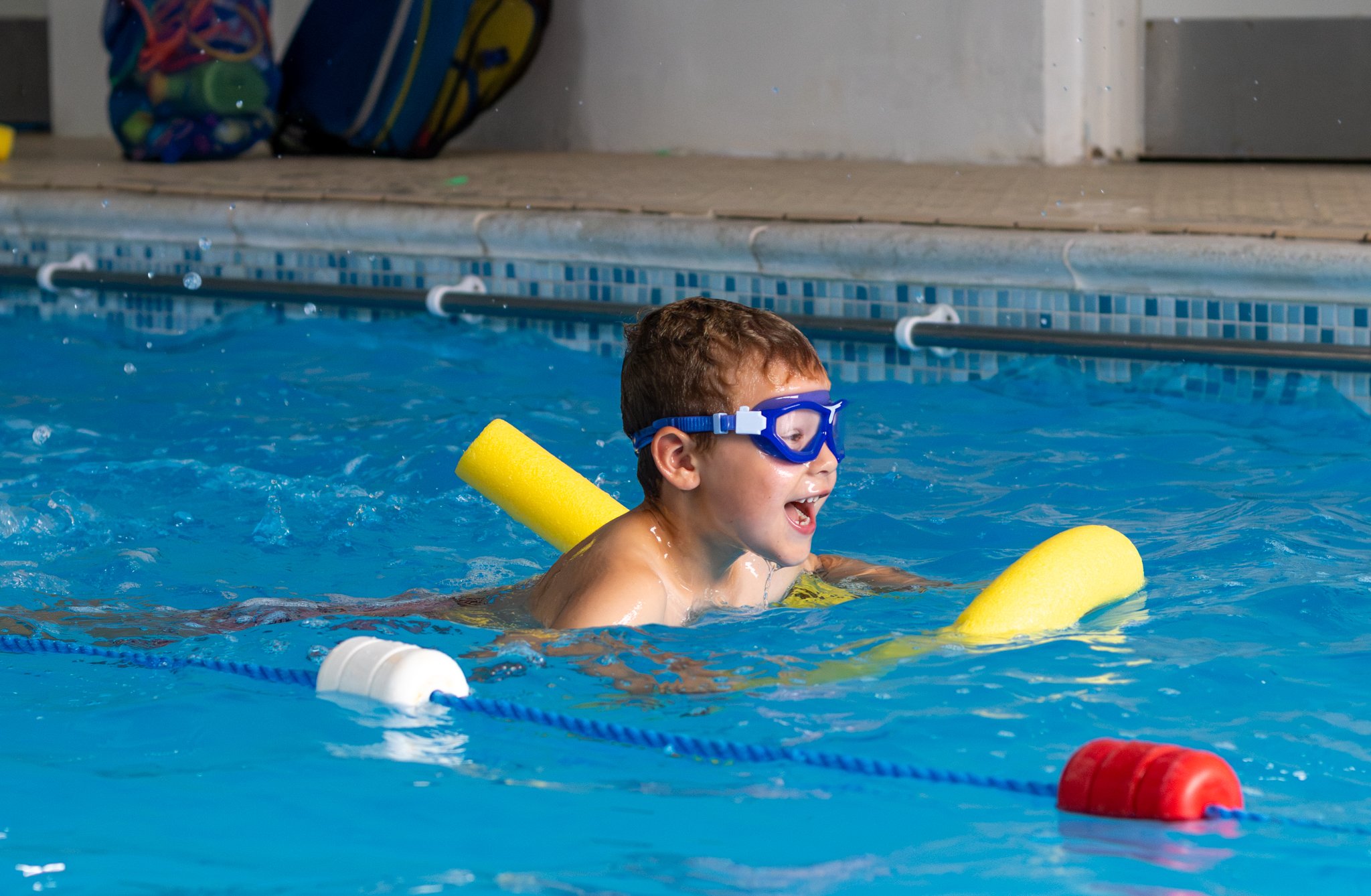 A boy swimming in a pool with a yellow pool noodle, wearing blue goggles, and smiling.