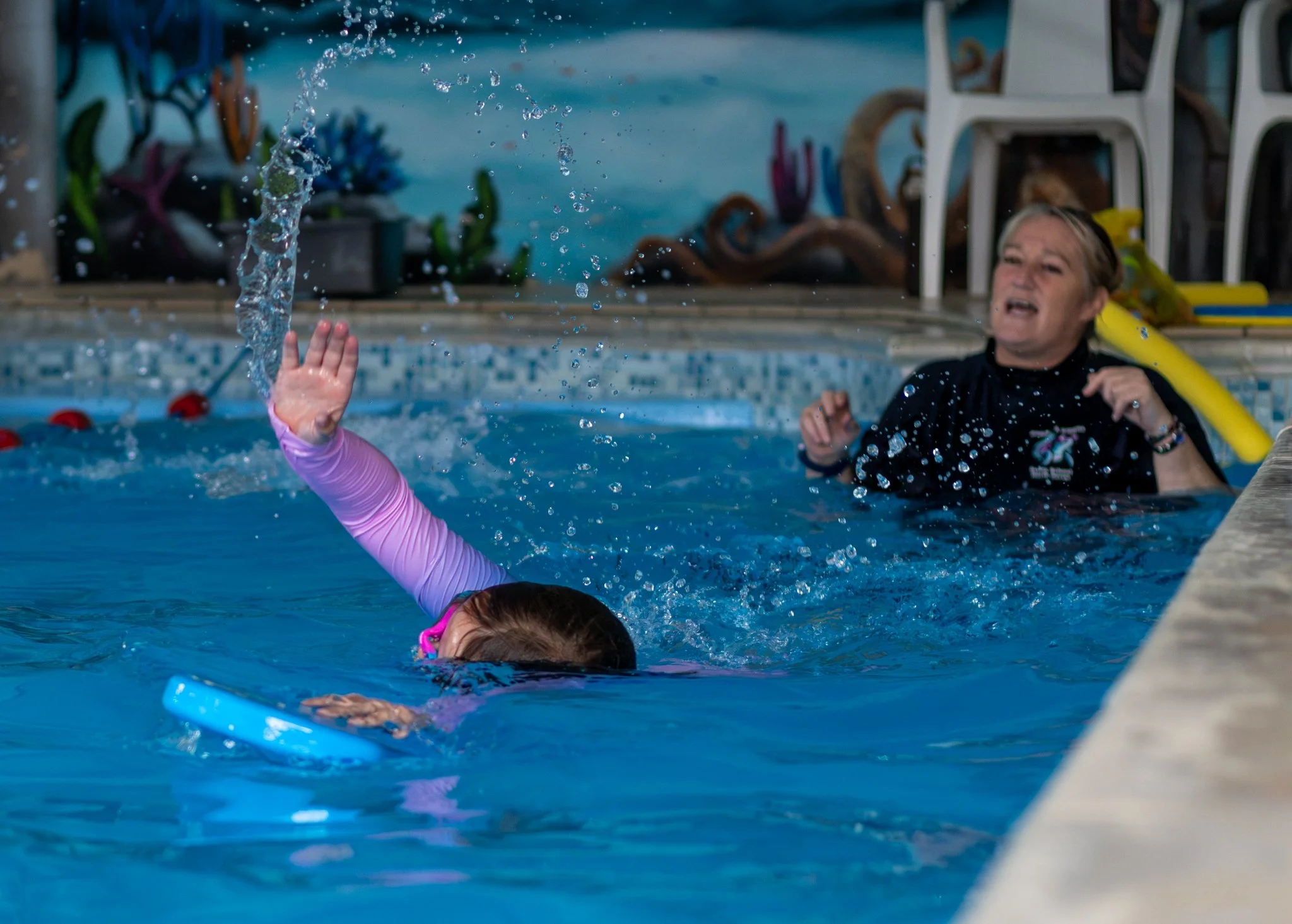 A young girl with pink swimming goggles and a pink long-sleeve swimsuit in a swimming pool, reaching up with her left hand as water splash from her hand, with a woman in a black swim shirt and blonde hair, smiling in the background, near bubble-filled pool toys and white plastic chairs.