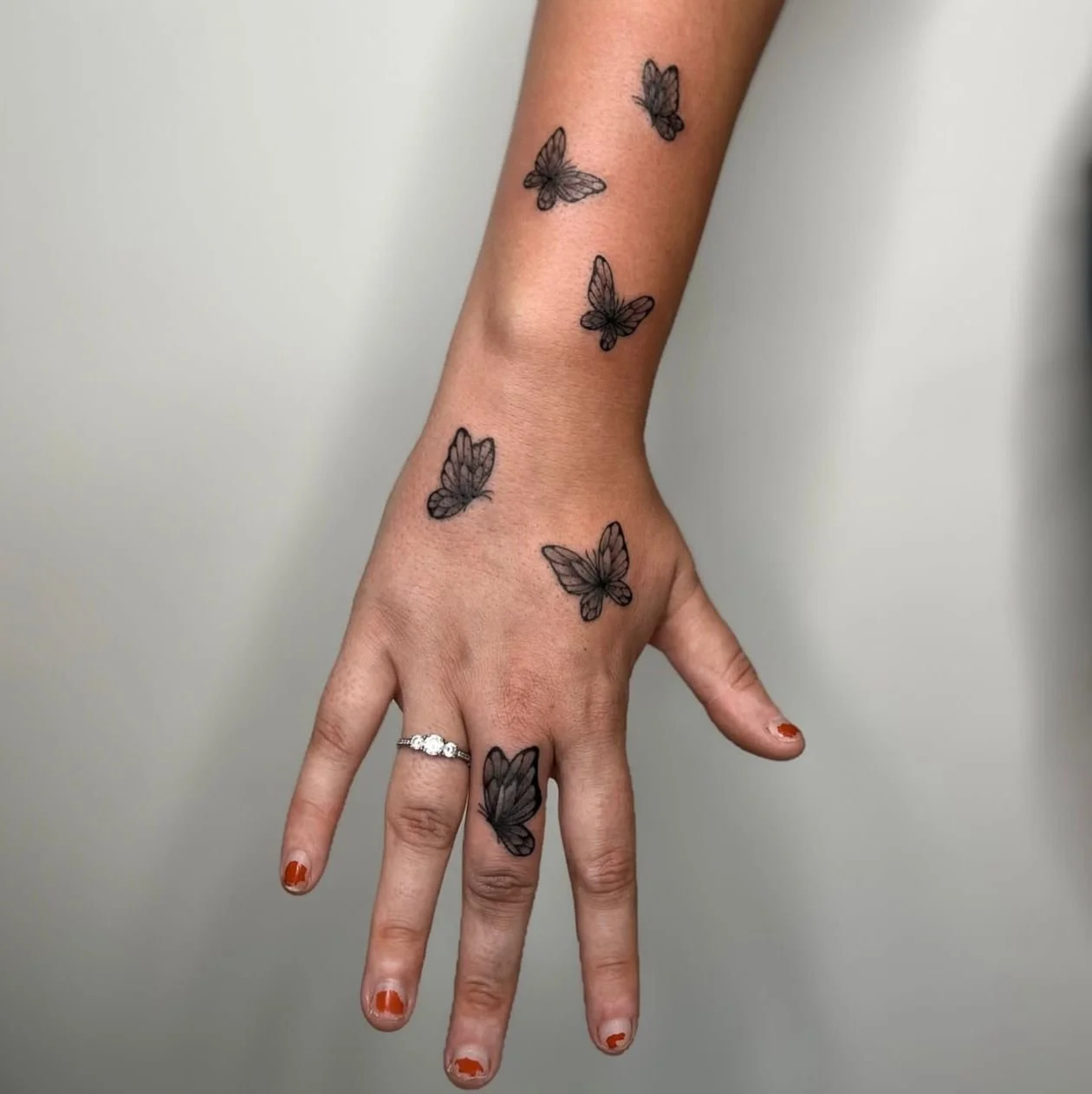 Dark black and grey tattoo of six small butterflies in total reaching from one on the middle finger, two butterflies on the back of the hand, and three on the lower wrist.