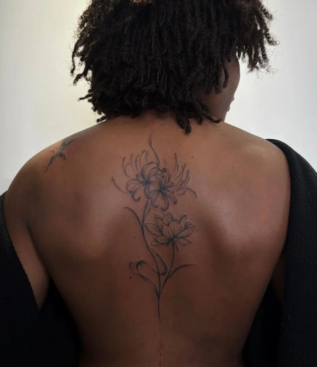 Black and grey wispy fineline style tattoo on a woman's spine featuring a fire lily and water lily with gentle shading.