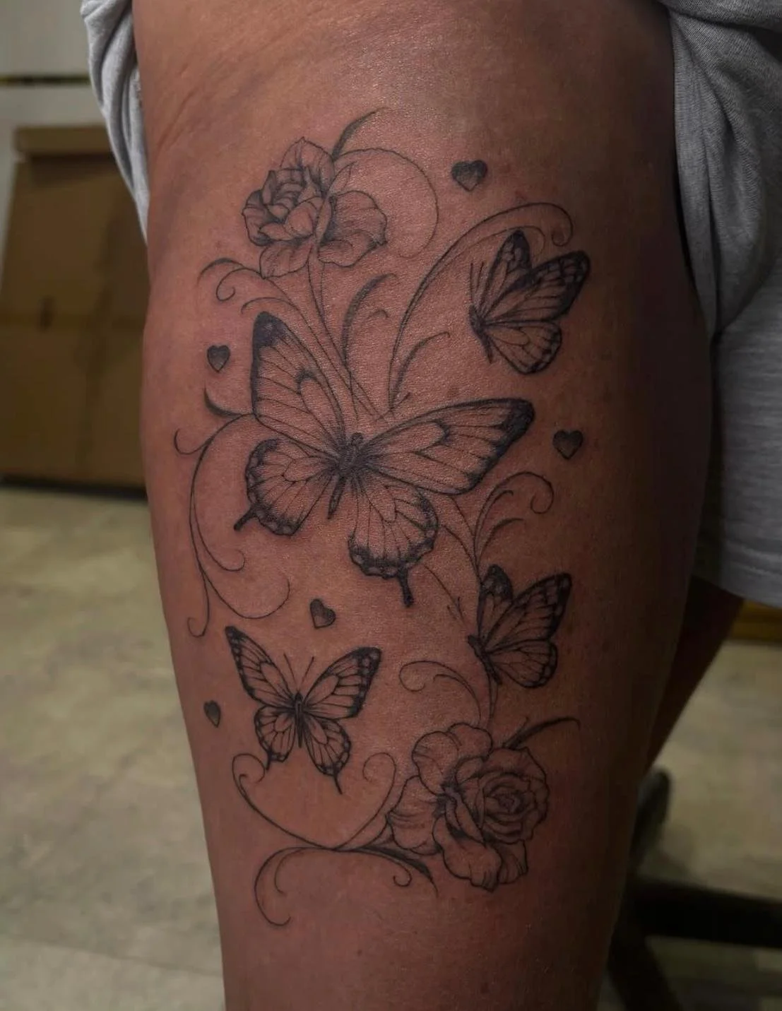 Thigh scaled black and grey tattoo featuring four butterflies surrounded by wispy detailing lines, hearts, and a rose at the top and bottom of the design.