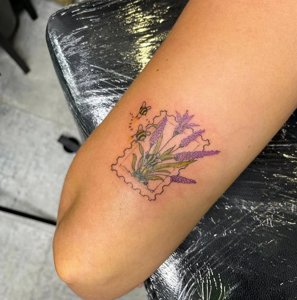 Delicate fineline and fine detailed colour tattoo of a postage stamp, featuring lavender reaching outside of the stamp, attracting two tiny bees. 