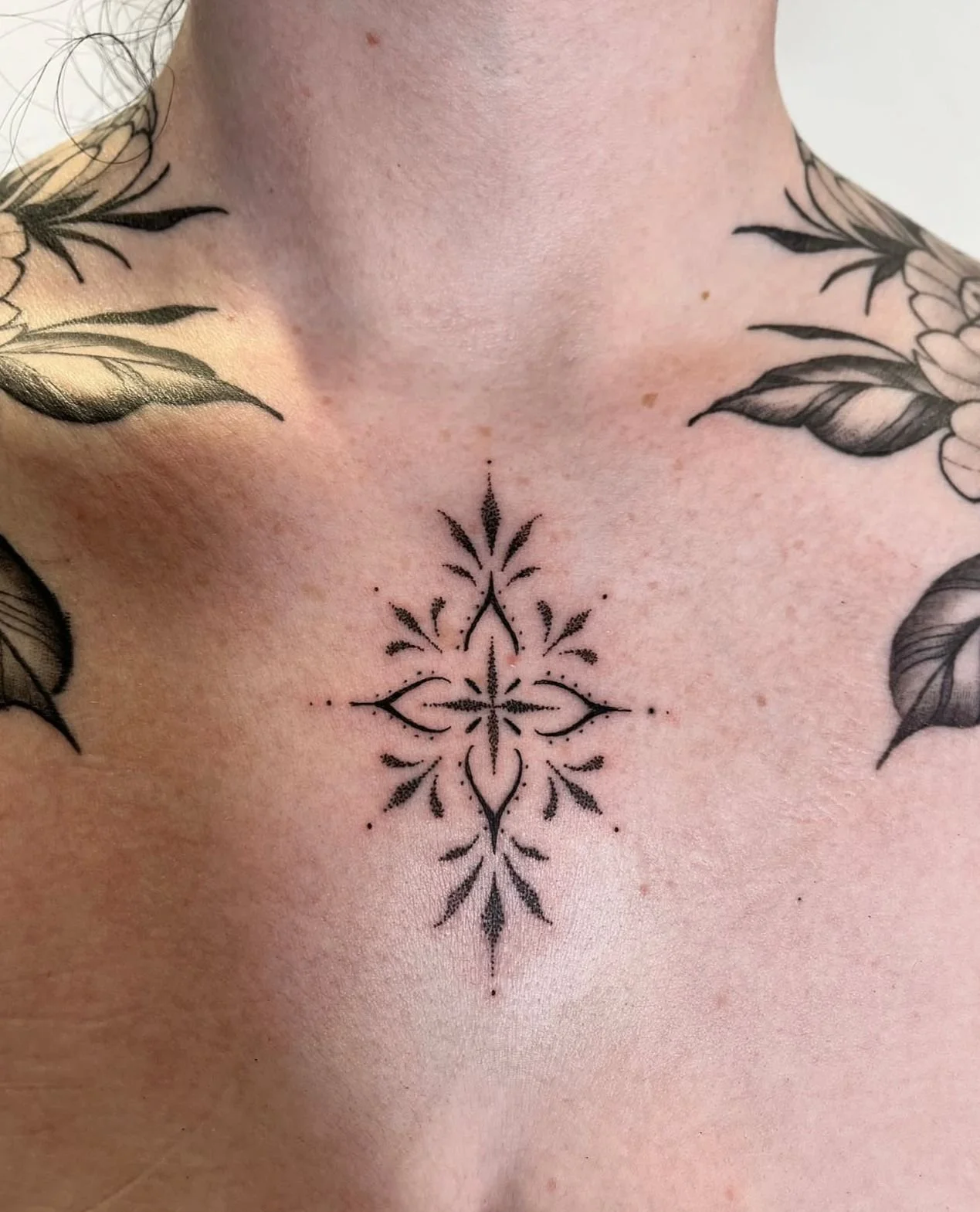 Black ornamental tattoo on a woman's chest between the collarbones, with dark leaf tattoos reaching on the chest from the shoulders. 