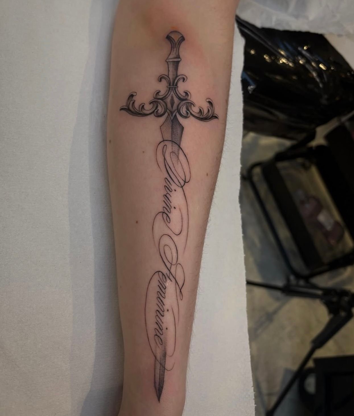 Fineline black and grey tattoo on a forearm of a sword, with 'Divine Feminine' written in script in place of the body of the blade.