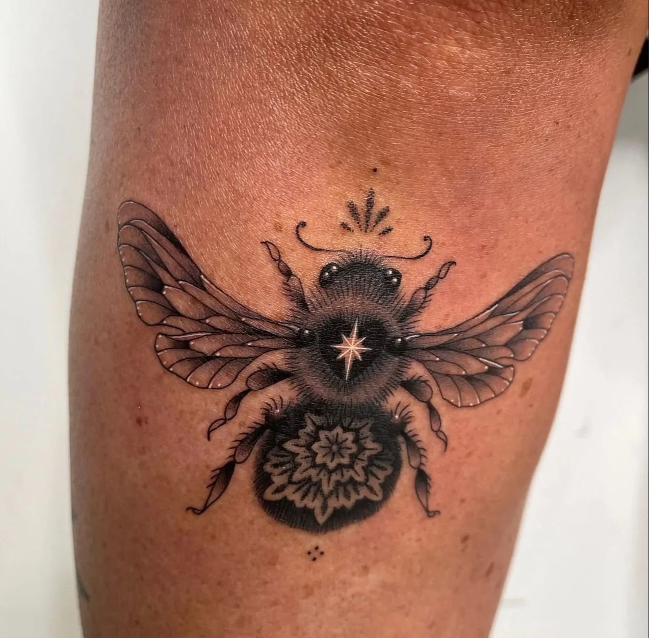 Dark black and grey tattoo of a bee with ornamental detailing, featuring a mandala, white detailed star, and other ornamental details.