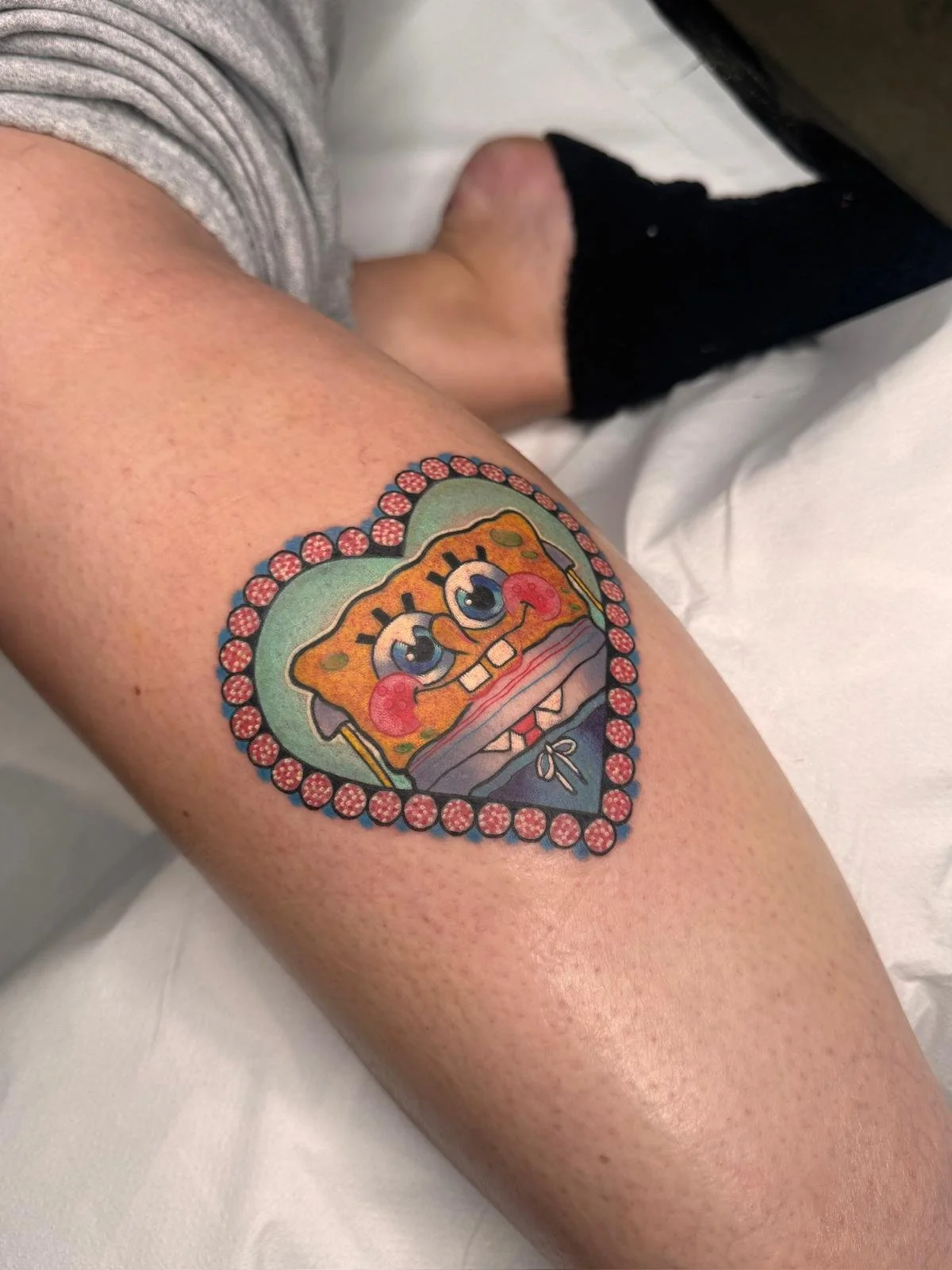 Neotraditional bold colour heart shaped tattoo on the side of the calf, featuring an ornamental beaded heart frame around the face of the charecter Spongebob Squarepants showing a cheeky cute expression.